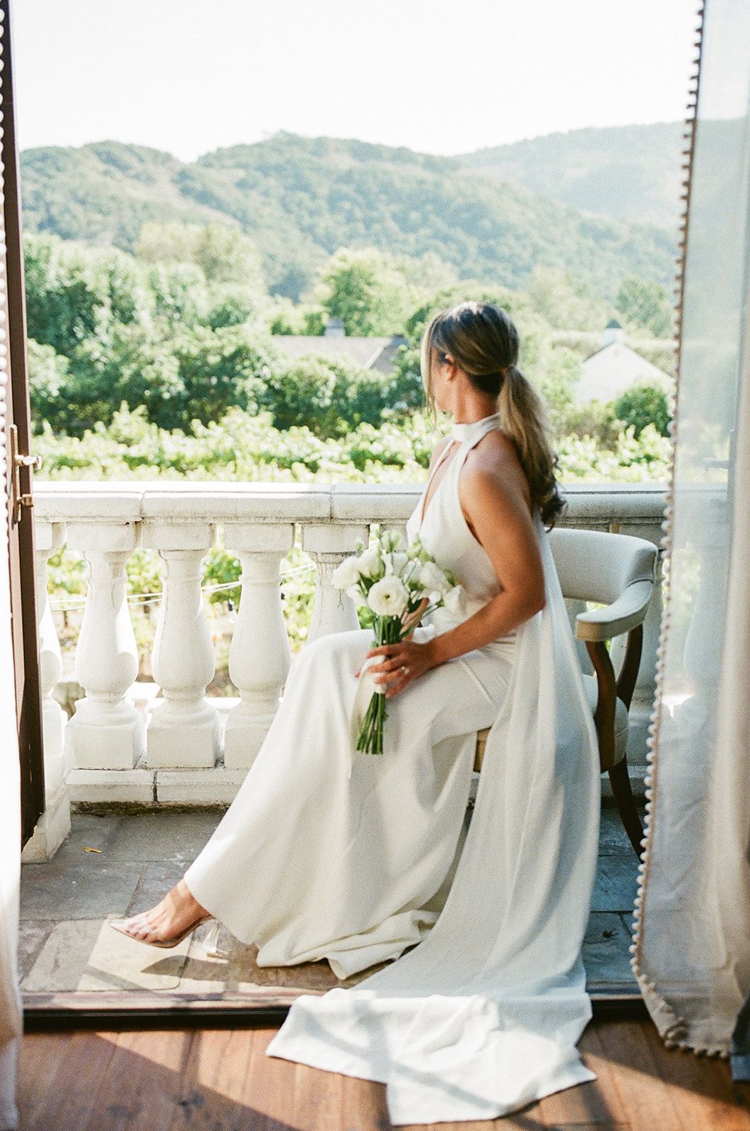 A bride in an elegant white gown holds a bouquet while sitting on a balcony overlooking mountain scenery.
