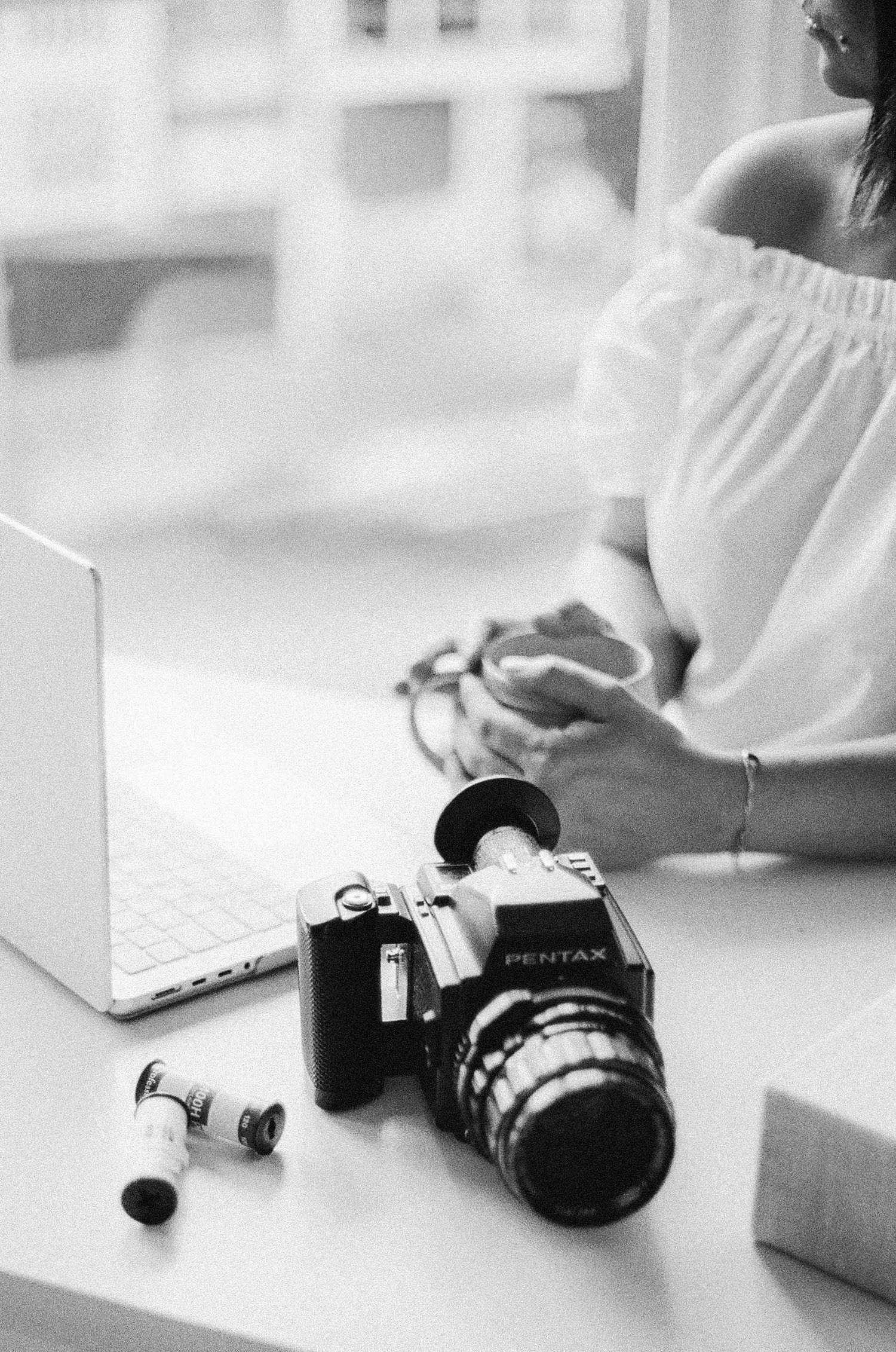 Black and white photo of a Pentax film camera with rolls on a desk beside a laptop and woman in white off-shoulder top.