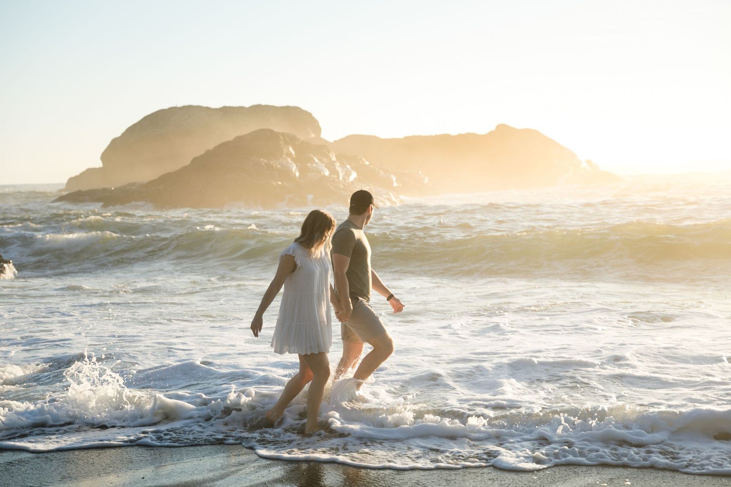 Couple playing in ocean waves during golden hour with rocky cliffs in background.