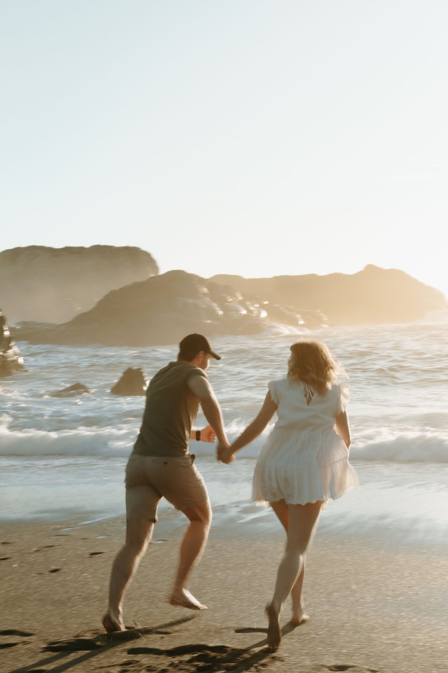 Playful couple running along beach shore during sunset with coastal cliffs behind them.