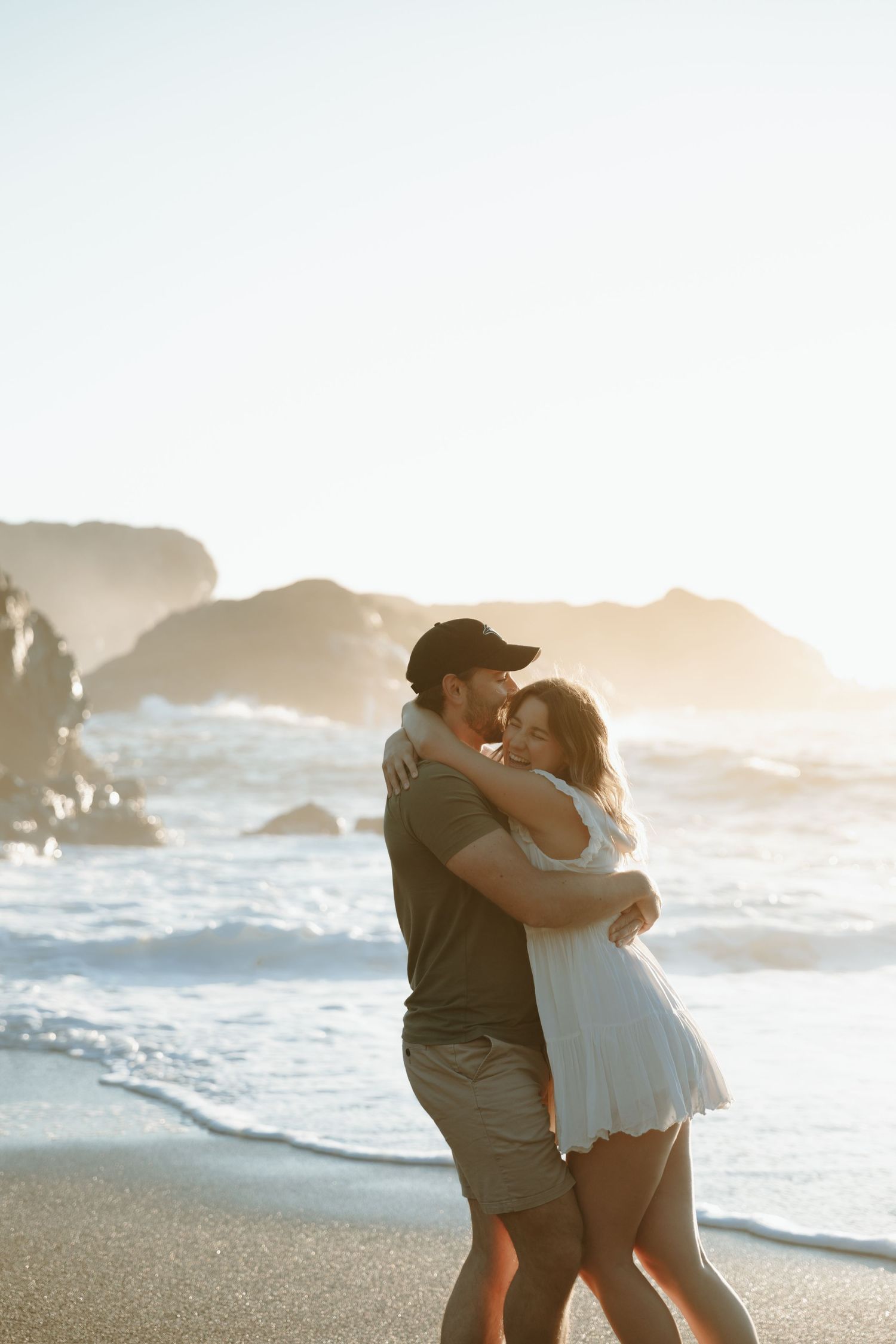 Intimate embrace on a beach at sunset with waves crashing against rocky coastline.