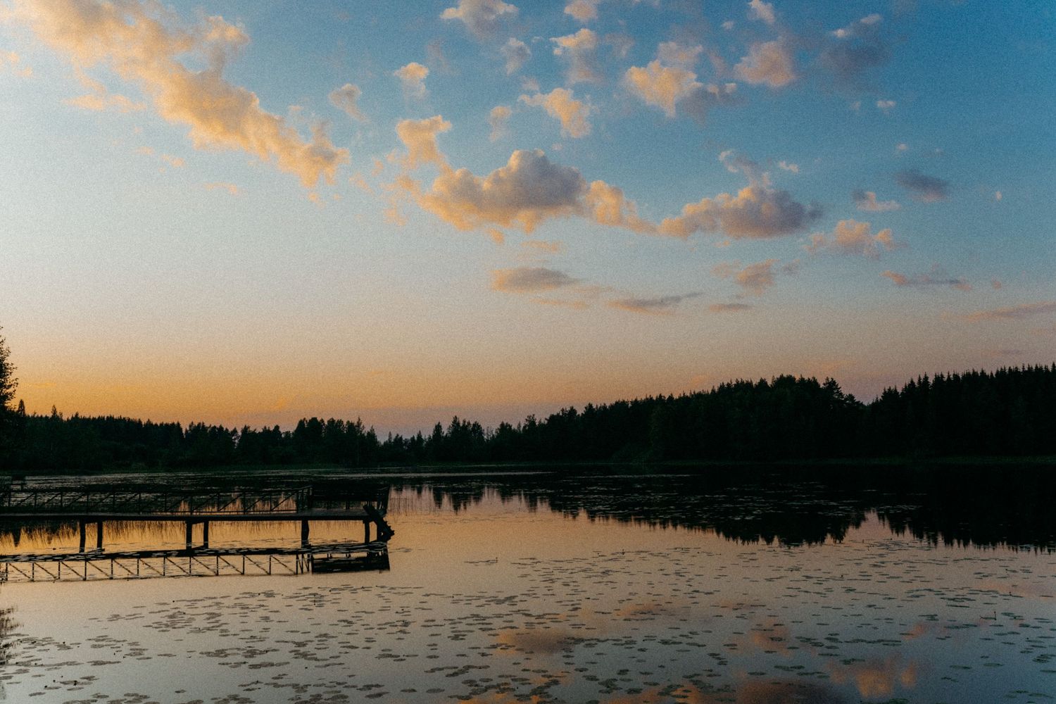 kesäinen mökkilaituri ja järvimaisema auringonlaskun aikaan. Pinkit pilvet heijastuu järven pinnasta.