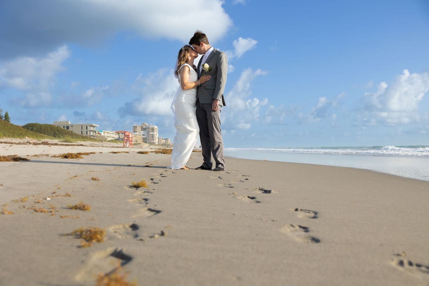 Newlyweds embrace on sandy beach with footprints trailing behind them.