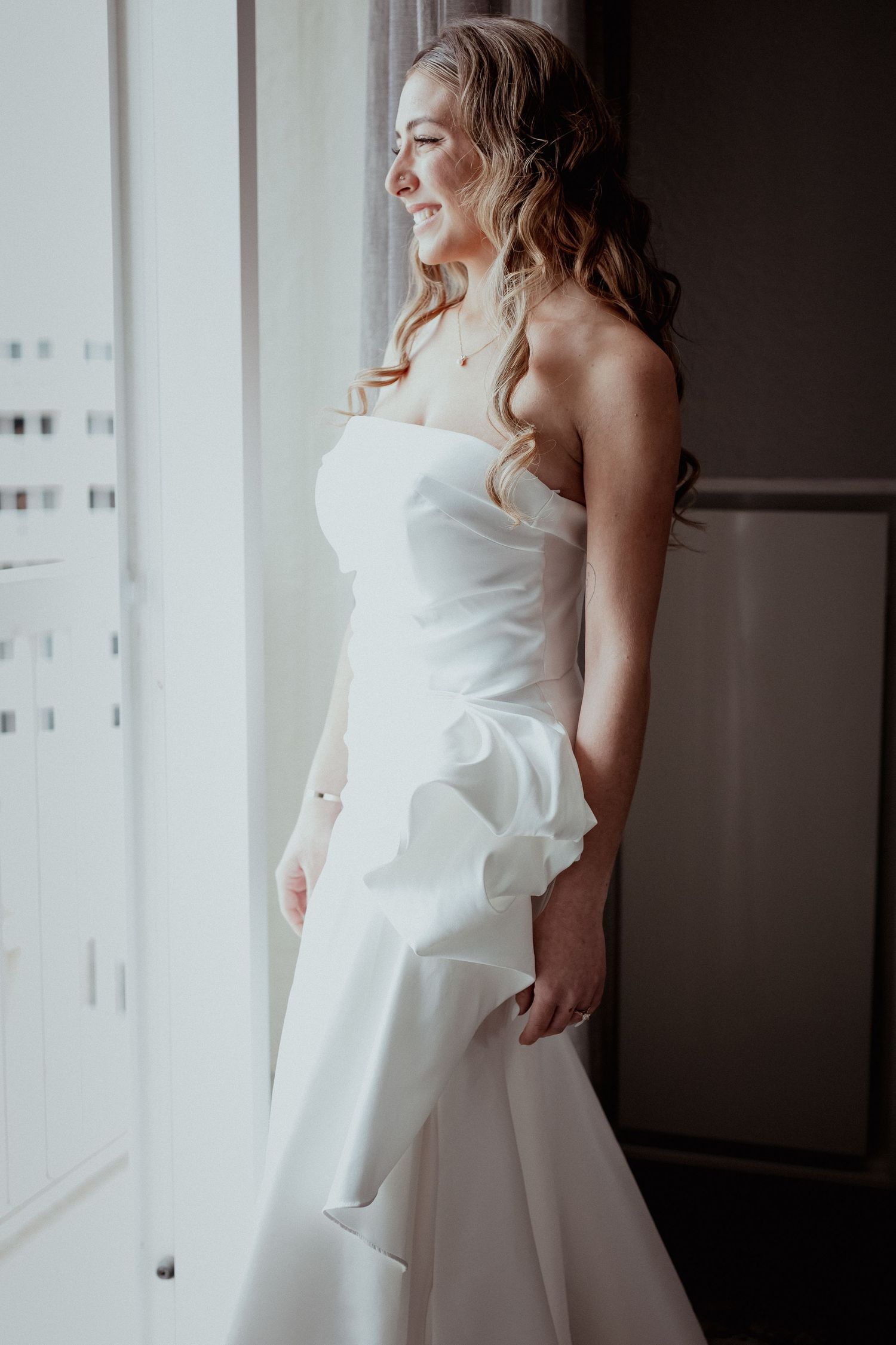 Candid bridal portrait by window showing elegant white wedding dress details.