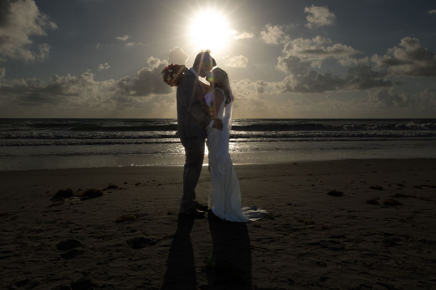 Silhouetted wedding couple embracing on beach at sunset with sun flare effect.