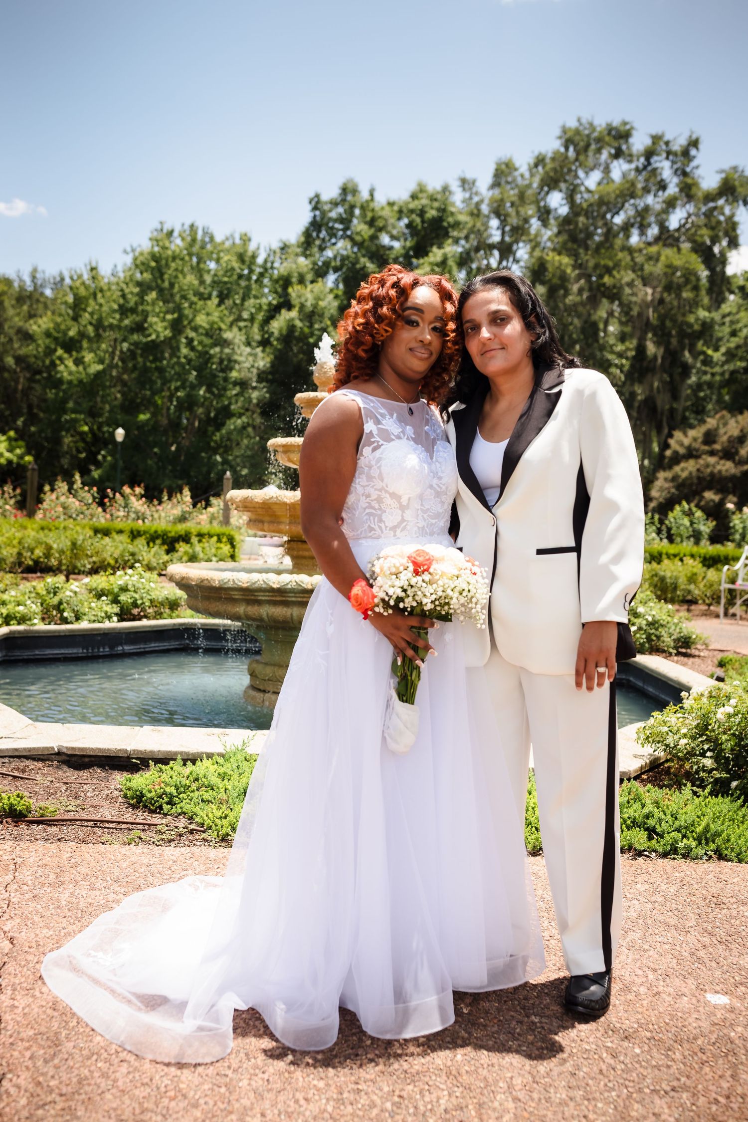 A wedding couple in white dress and white suit pose in front of a garden fountain on their special day.