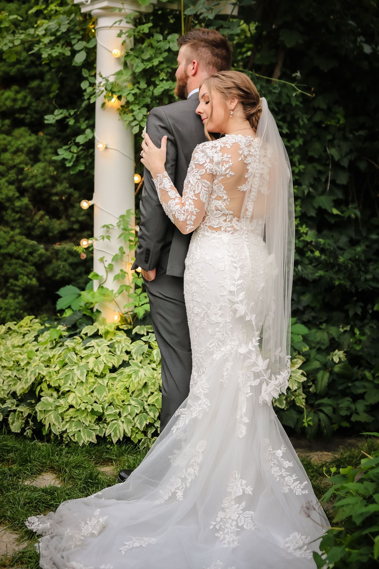 Bride in lace gown and groom share intimate moment against garden pillar with greenery.