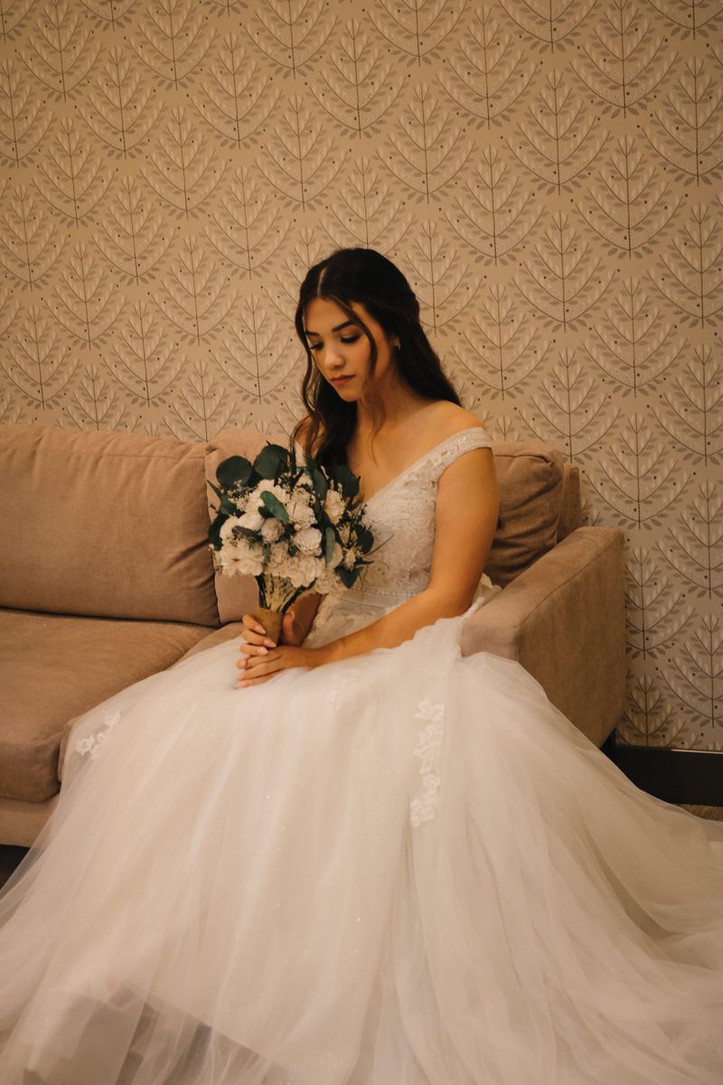 Bride in lace wedding dress sits on beige couch holding white and blue floral bouquet.
