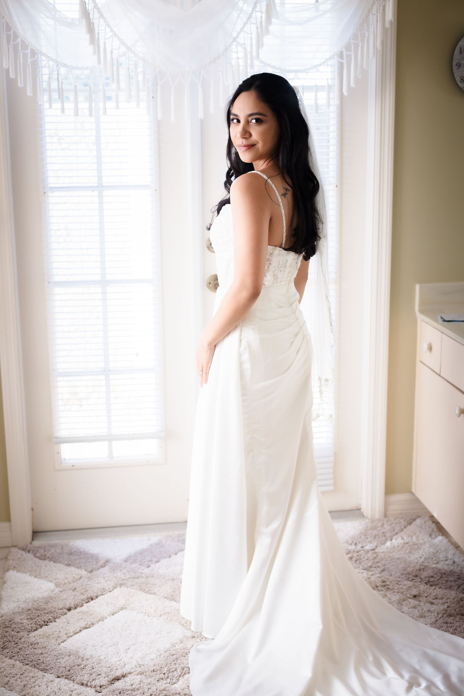 Bride in elegant white wedding gown poses gracefully by window with flowing white curtains and natural light.