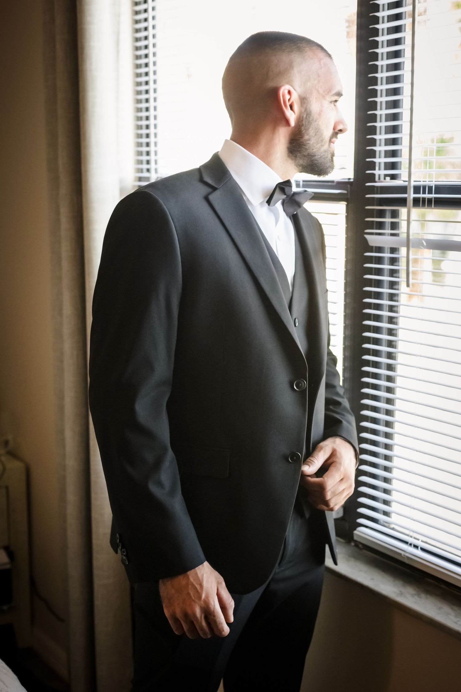 Man in dark suit gazing through window blinds in contemplative pose.