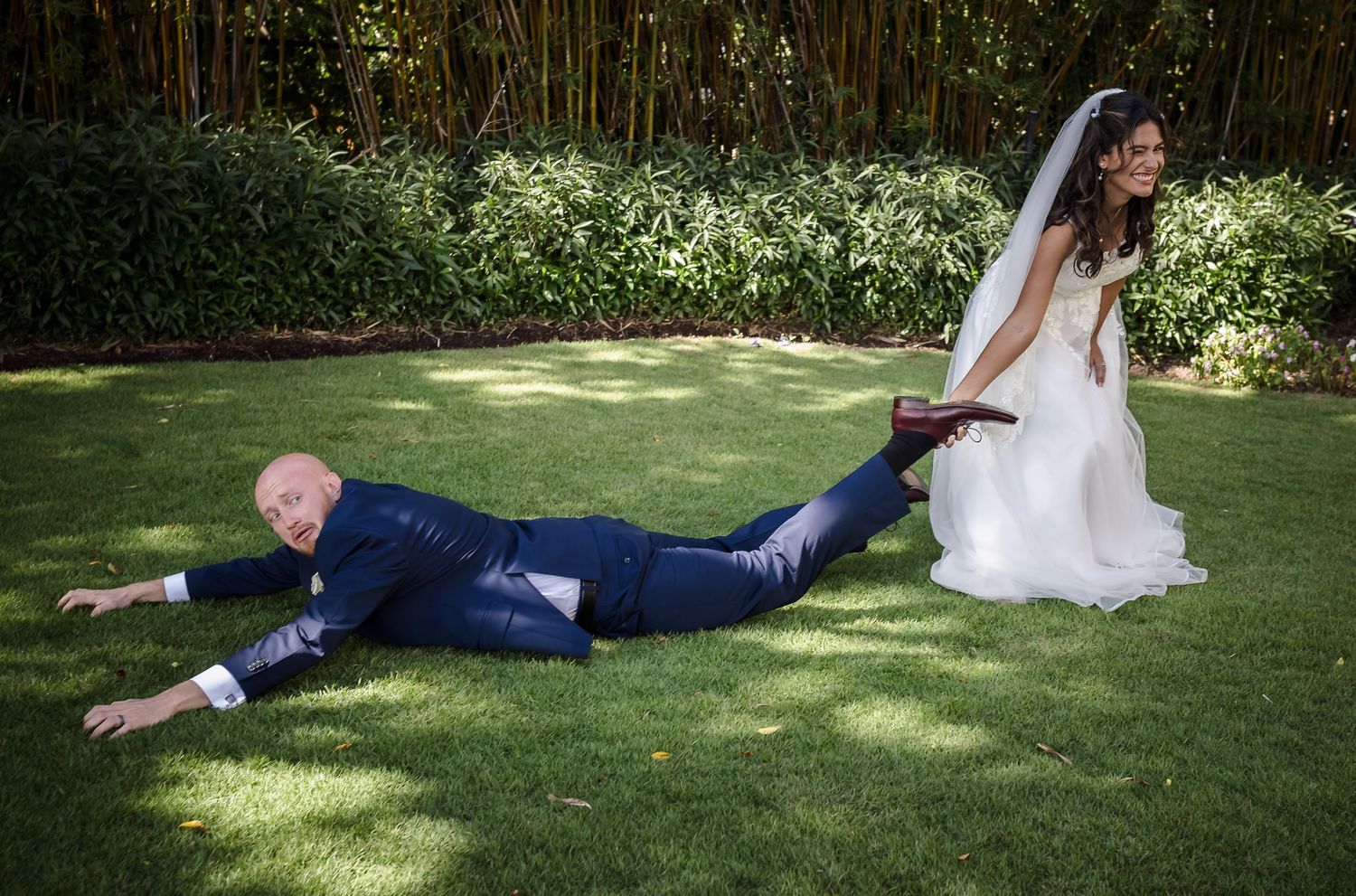 Humorous wedding photo with bride seated while groom lies on grass in garden setting.