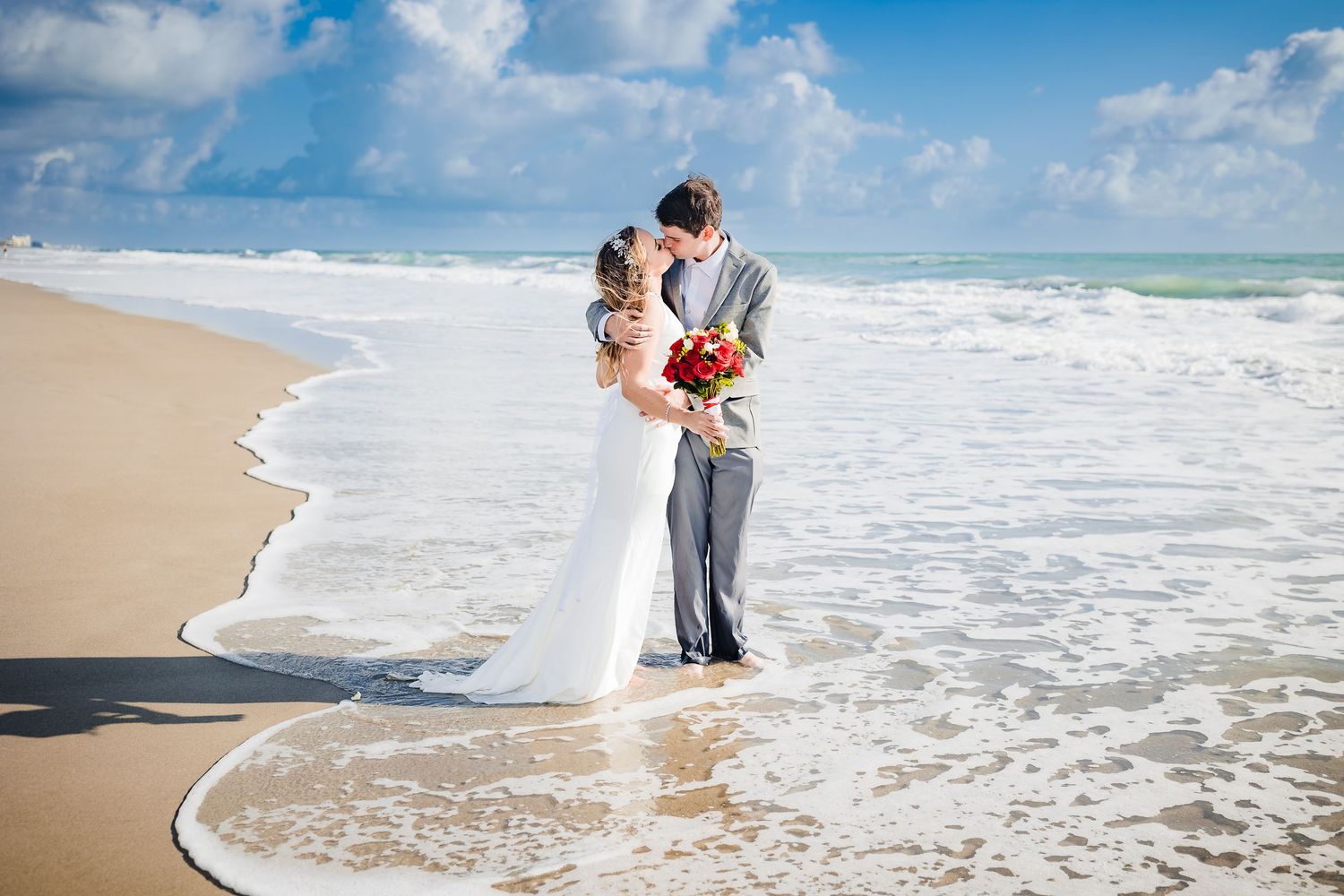 A romantic couple embraces on a sandy beach as ocean waves roll in under blue skies.
