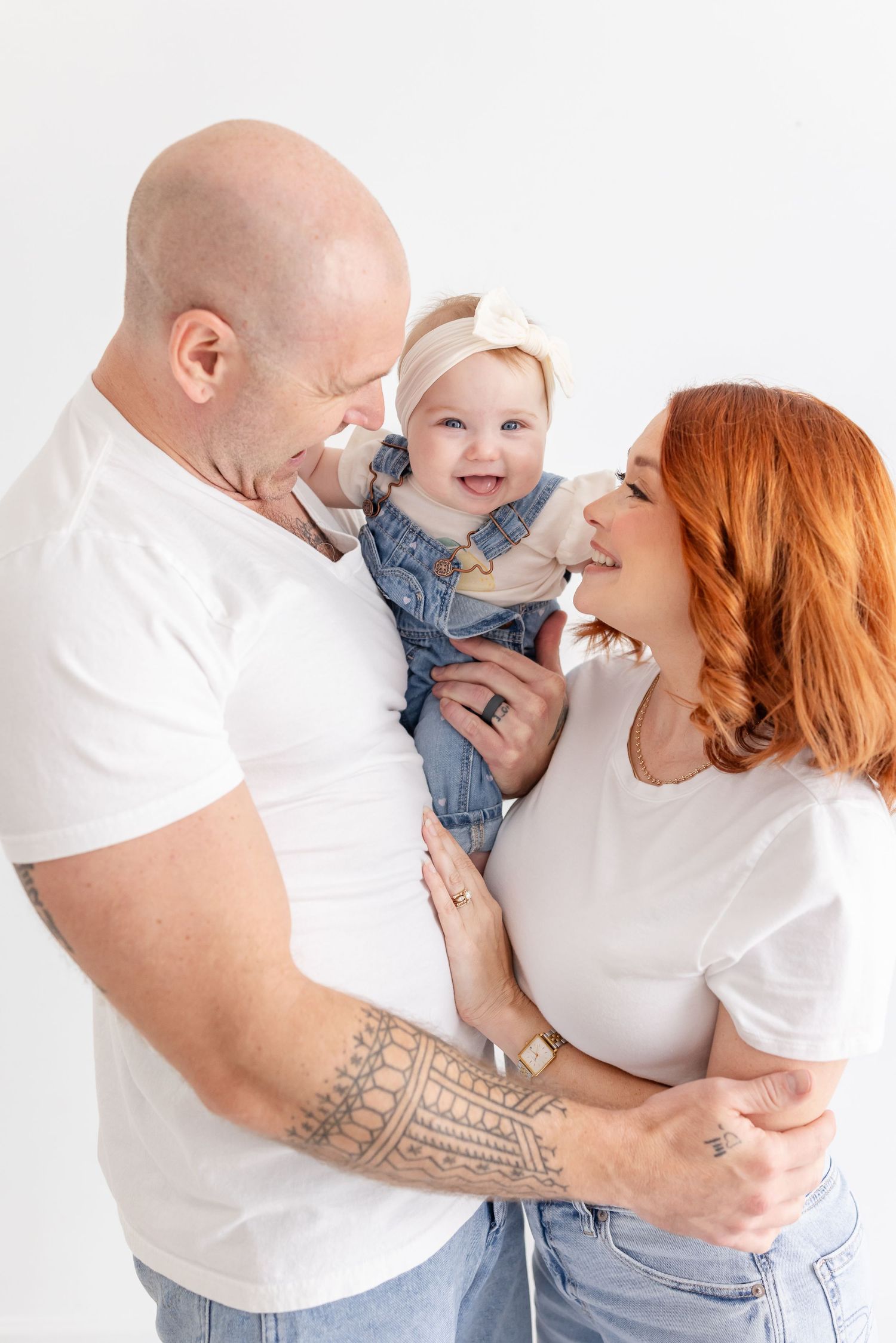 Happy family portrait with bald tattooed father, redhead mother in white tee, and smiling baby girl in denim outfit.