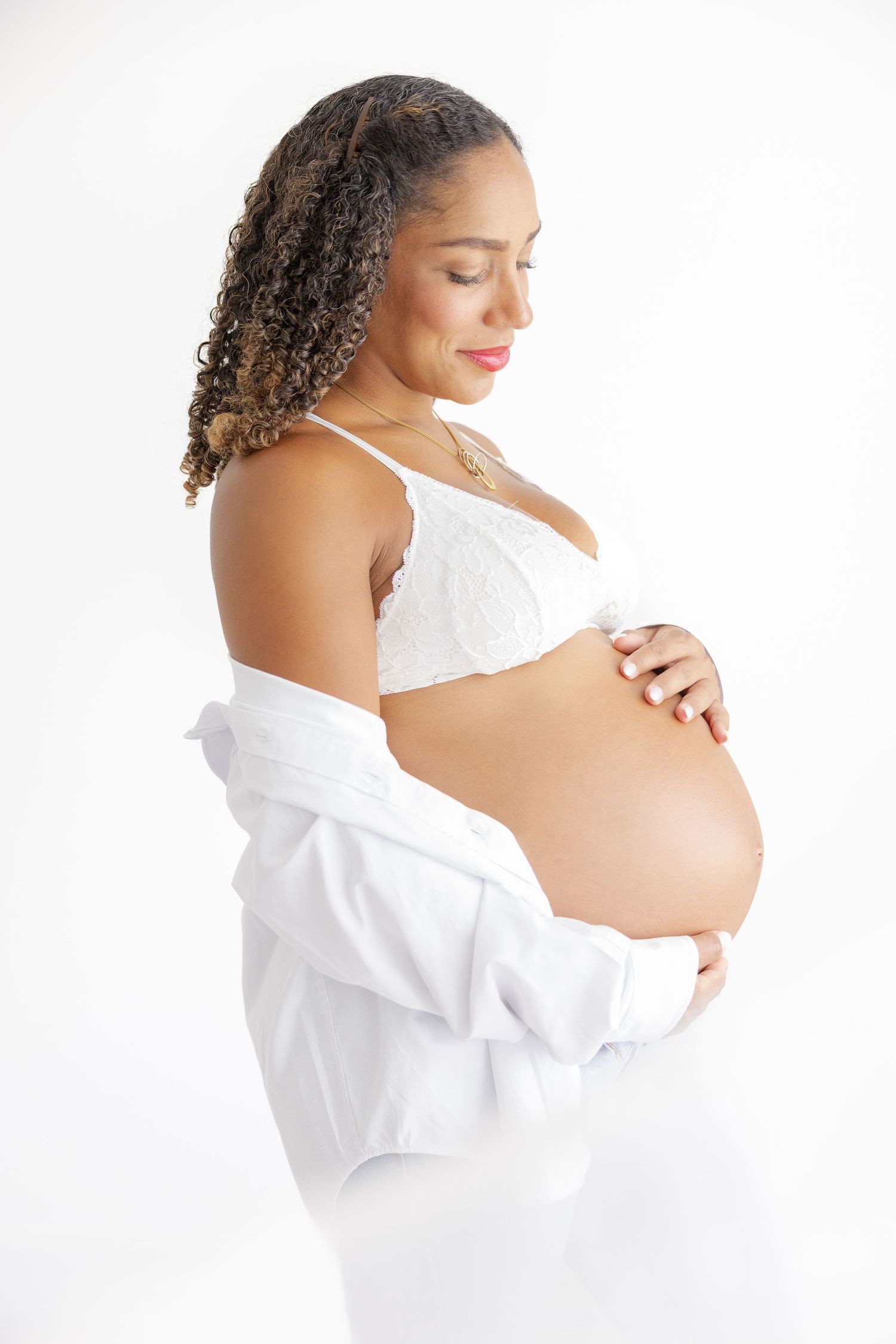 Pregnant woman in white bra and open shirt cradling her baby bump against a white background.
