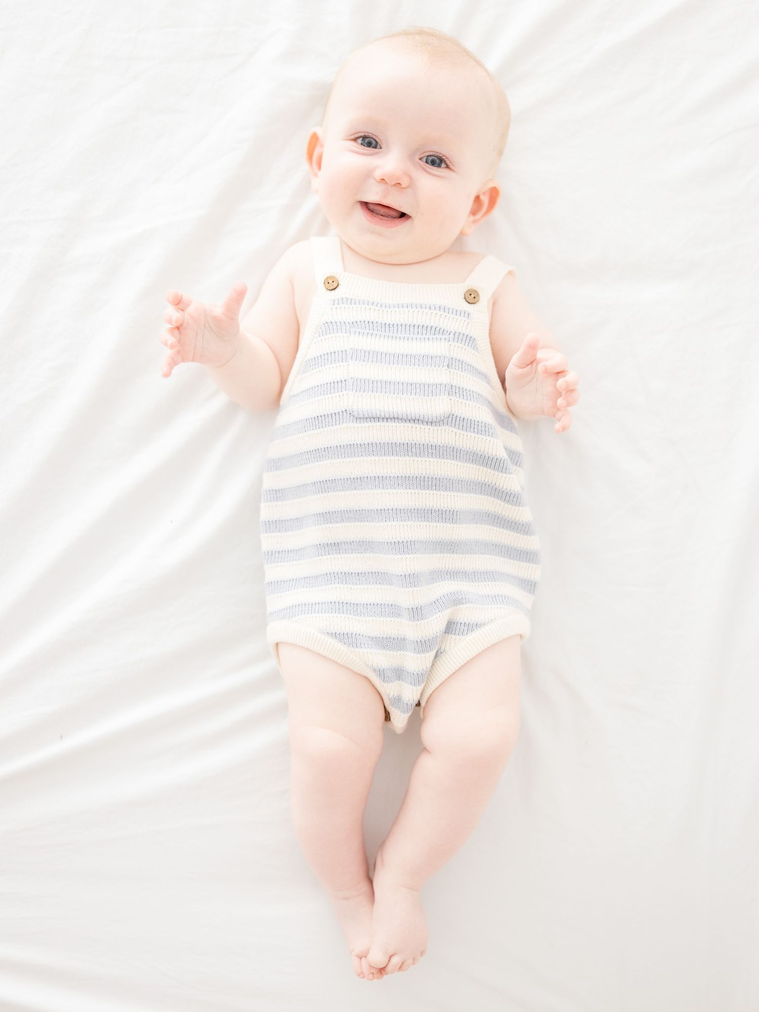 Smiling baby in a blue striped sleeveless romper lying on a white surface, looking up with bright blue eyes.
