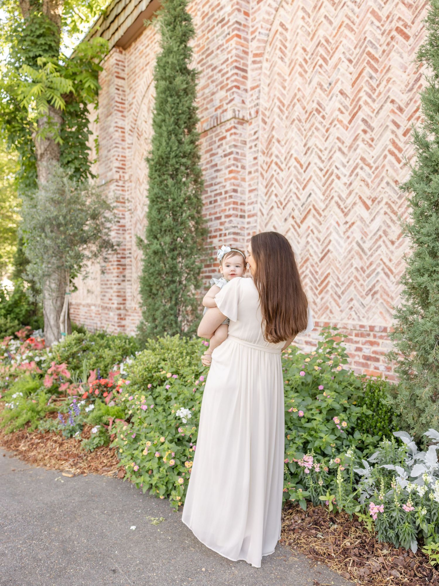 Mother in white dress holds baby girl near brick wall garden with colorful flowers and tall cypress trees.