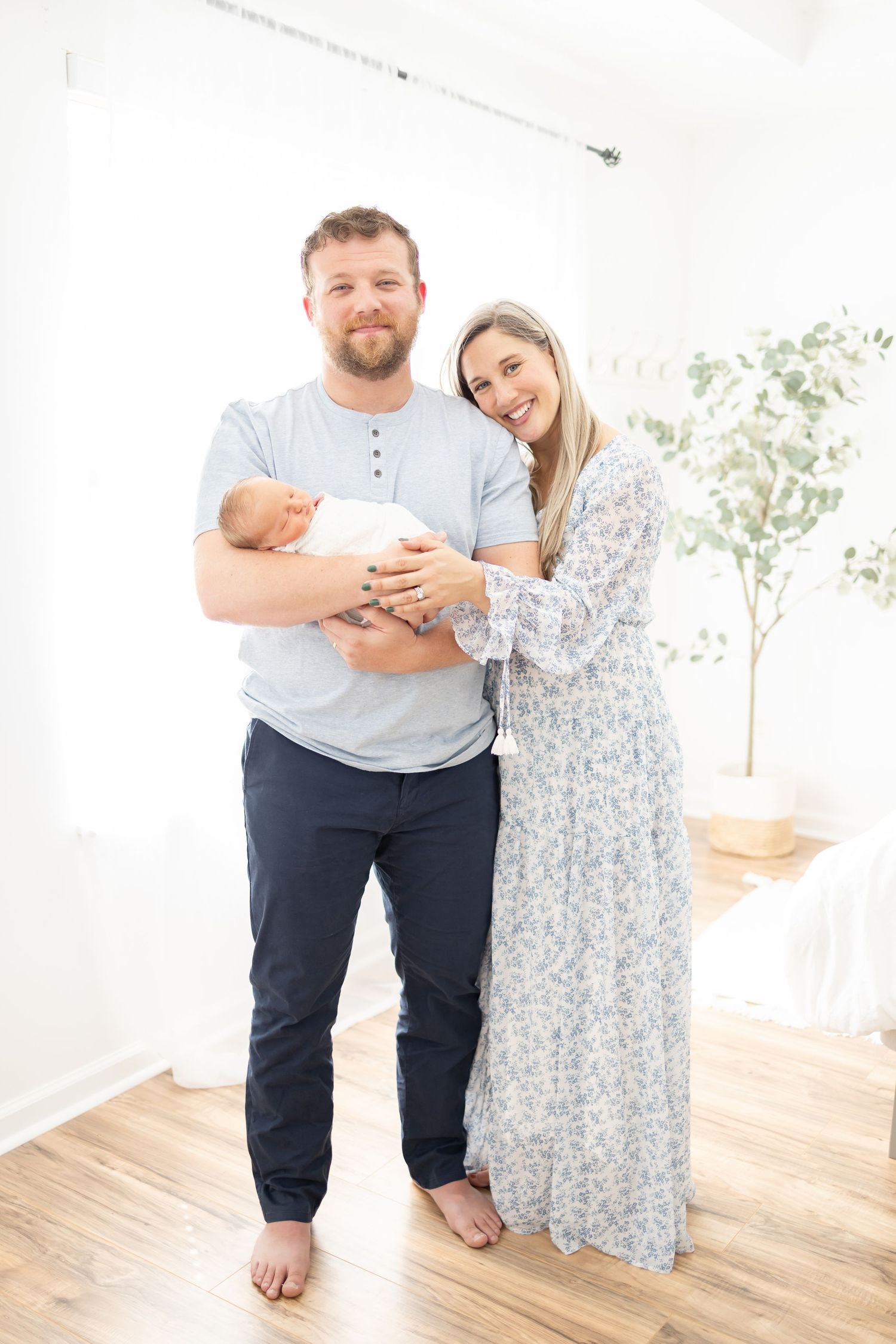 A couple stands together in a bright room holding their newborn baby during their Columbus, GA Newborn Portrait session.