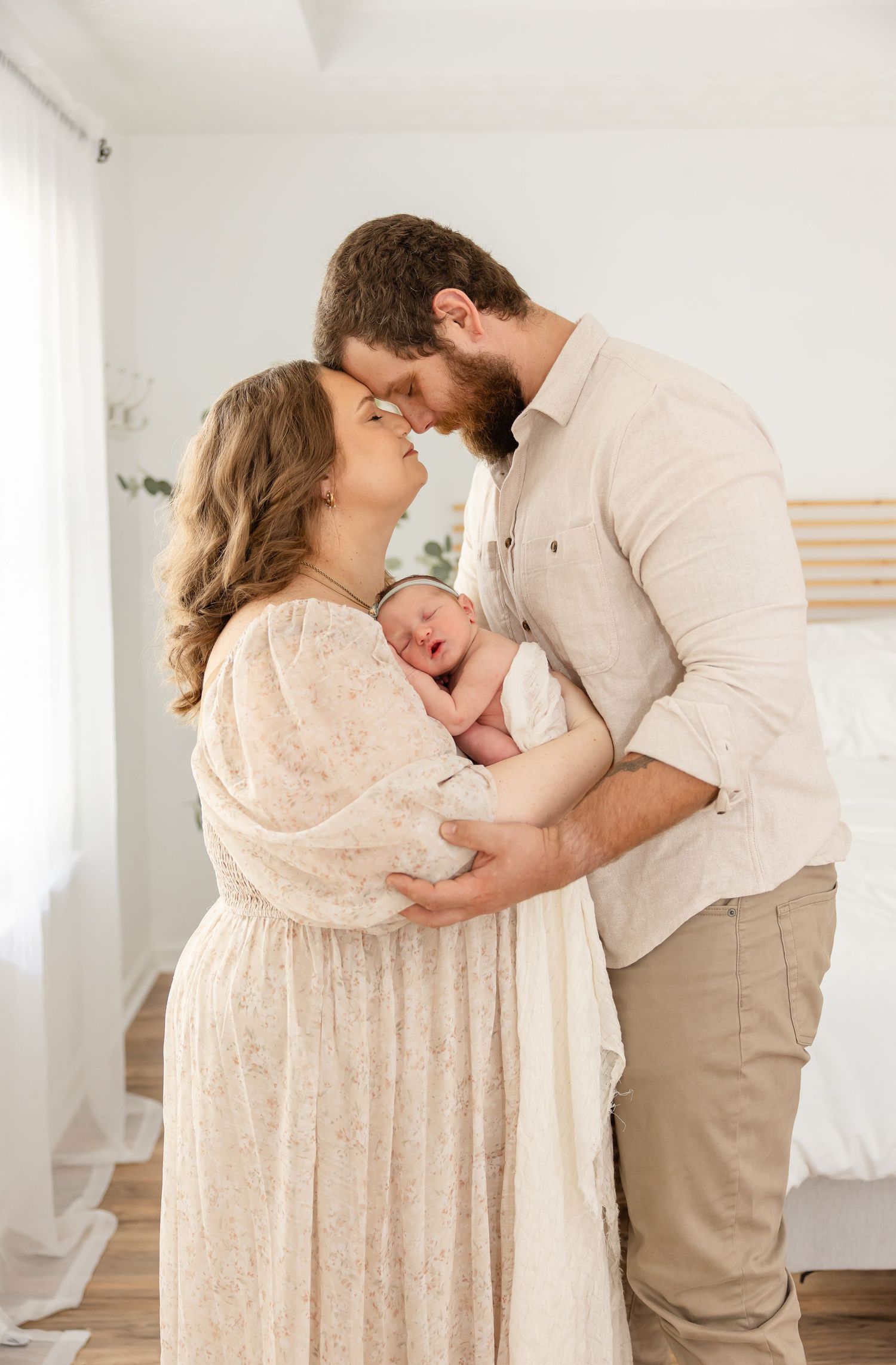 Loving couple in neutral tones cradle their newborn baby in a tender embrace against white walls.