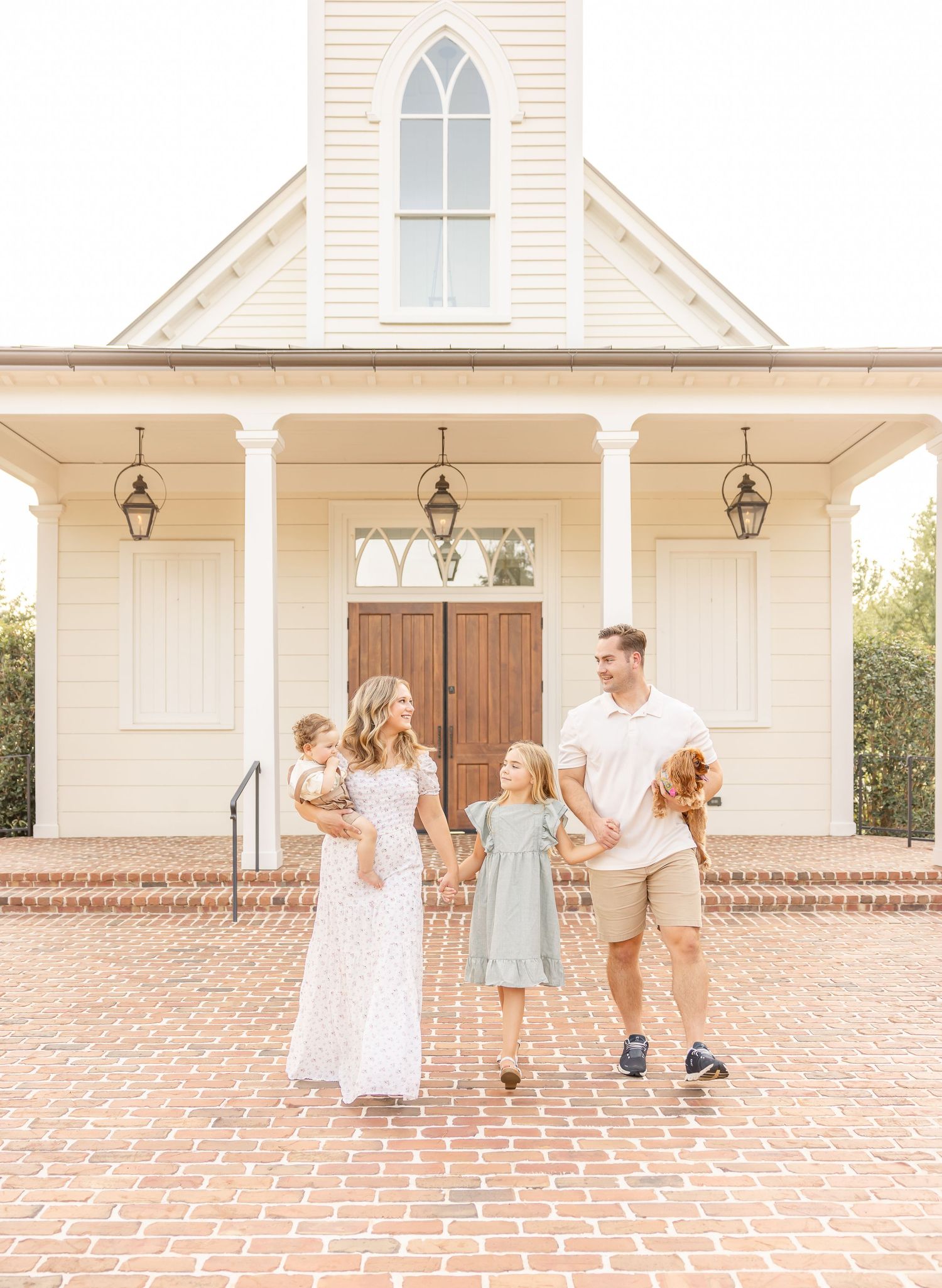 A family in coordinated light outfits stands together on a brick sidewalk in front a white church holding hands for their family photos.