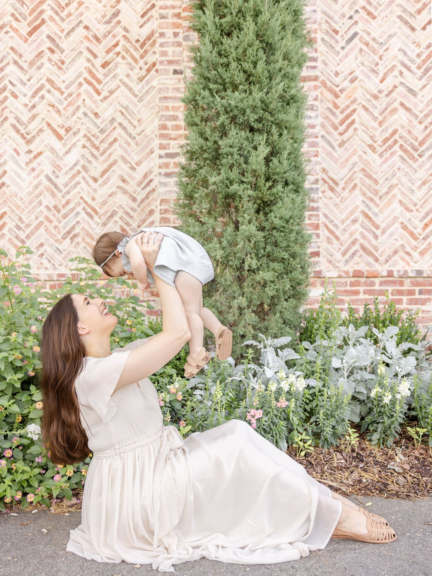 A mother sits on pavement lifting her baby in the air, smiling, in front of a brick wall with greenery.