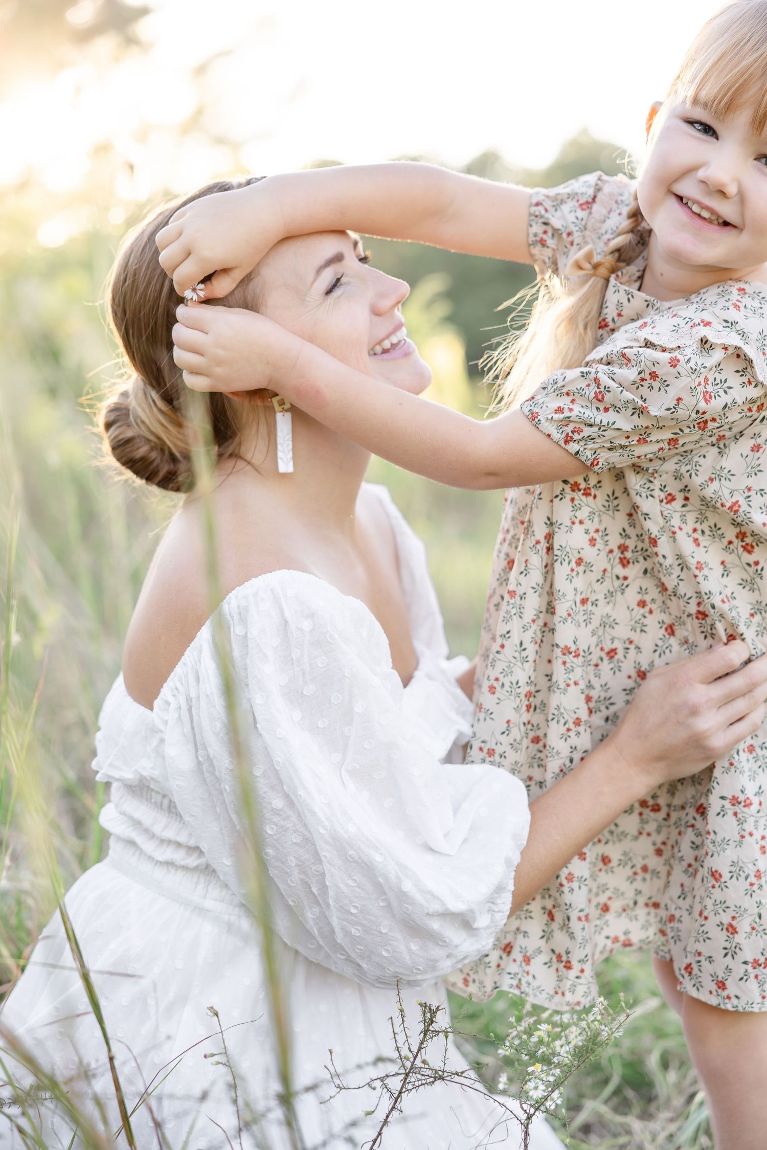 Smiling mother in a white dress and her young daughter in a floral dress share a playful moment in a sunlit grassy field.