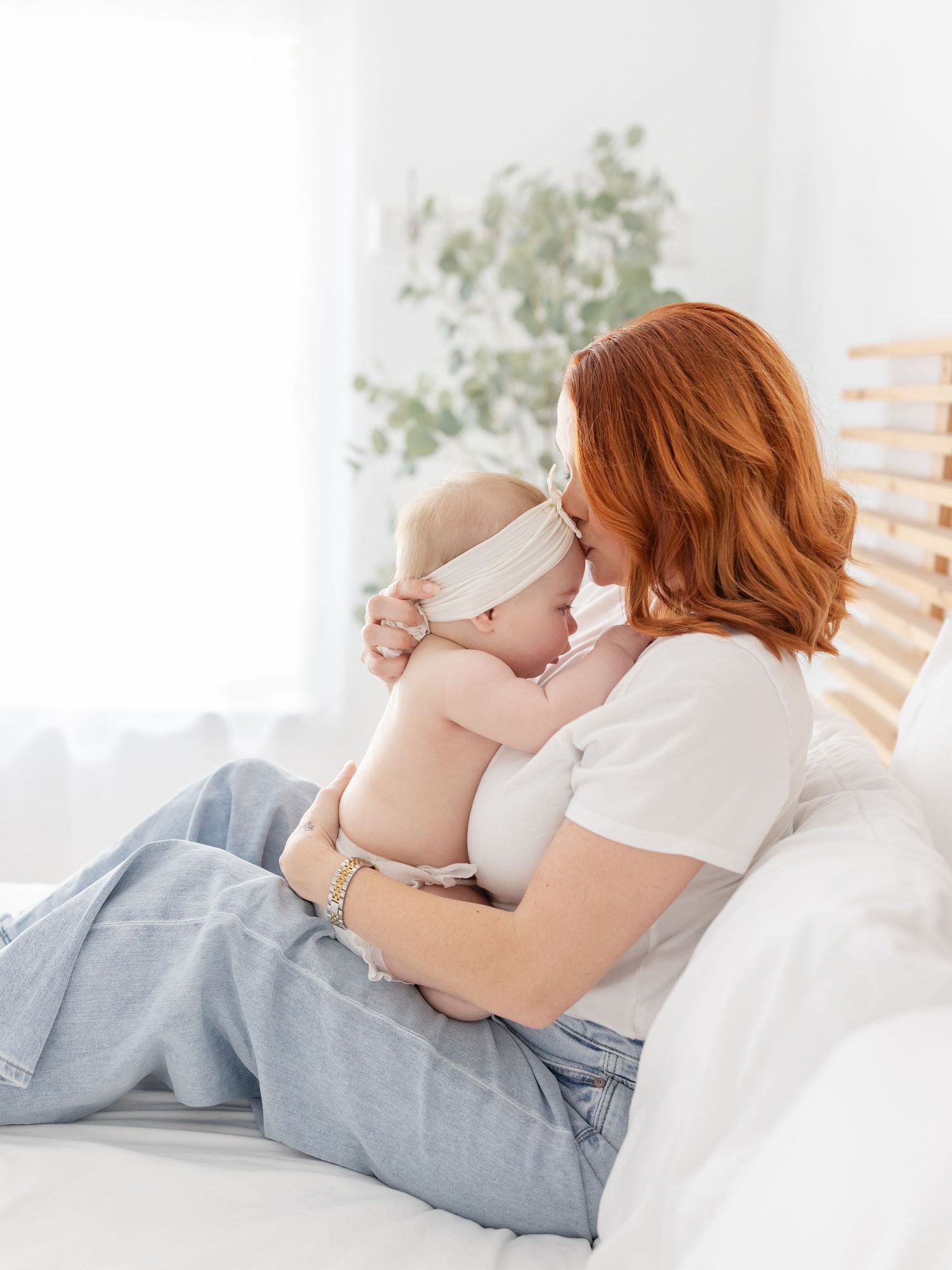 Redheaded mother tenderly holding her baby girl in a white bow headband while relaxing on a bed.