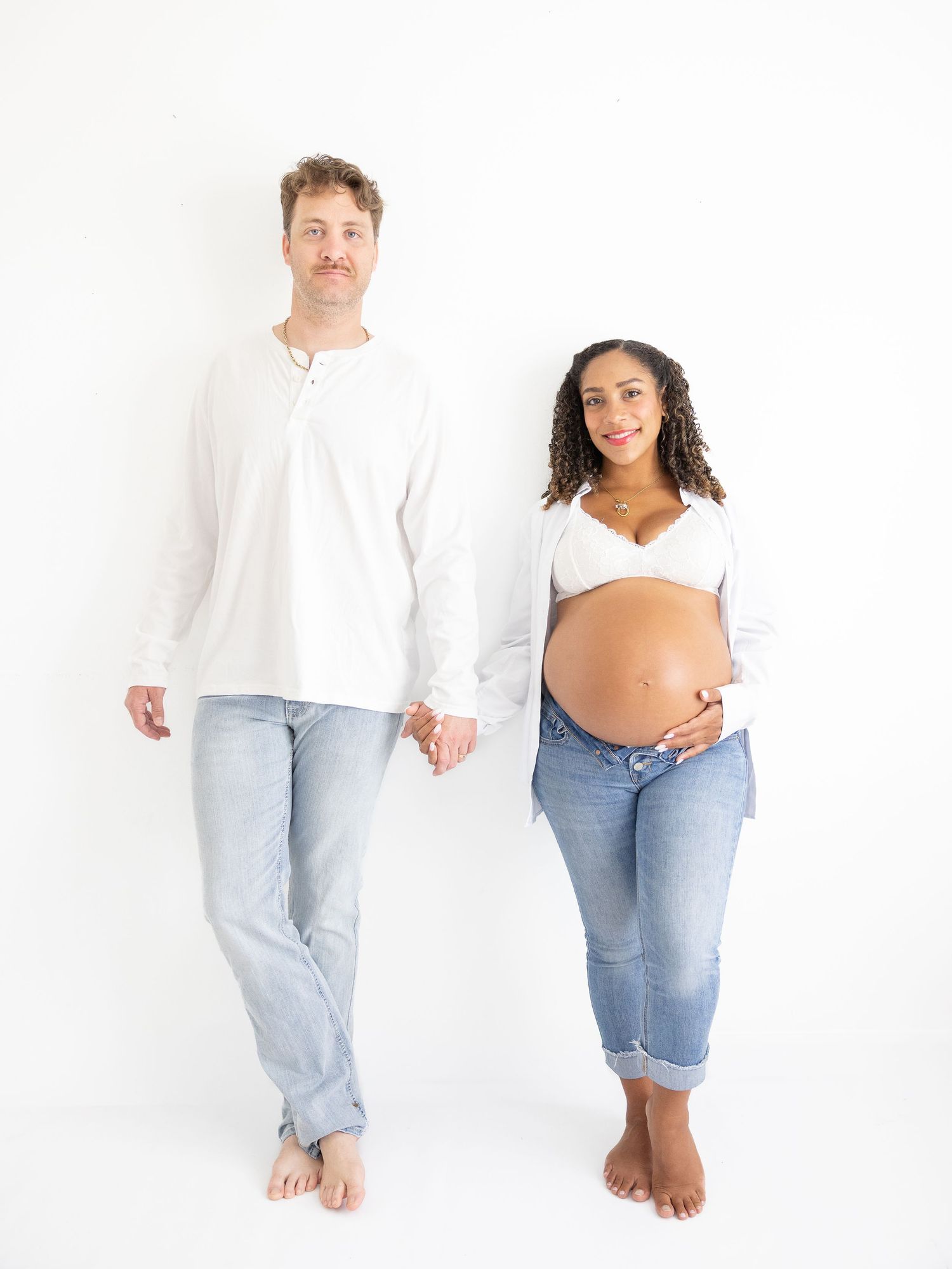 A couple in white tops and jeans pose together, the woman visibly pregnant, holding hands against a white background.
