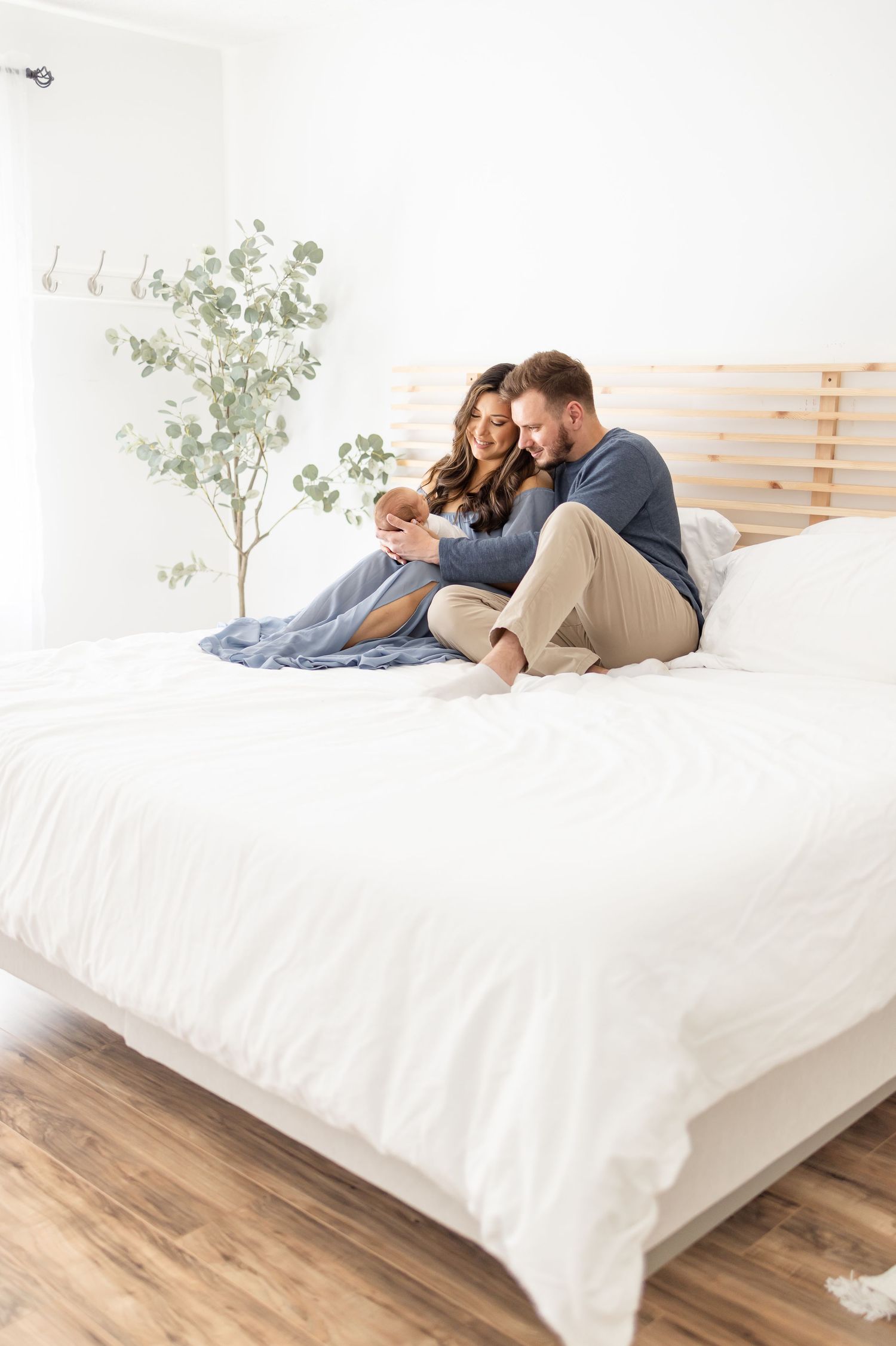A couple relaxes together on a white bed while gazing down at their newborn at their studio newborn session in Columbus, GA.