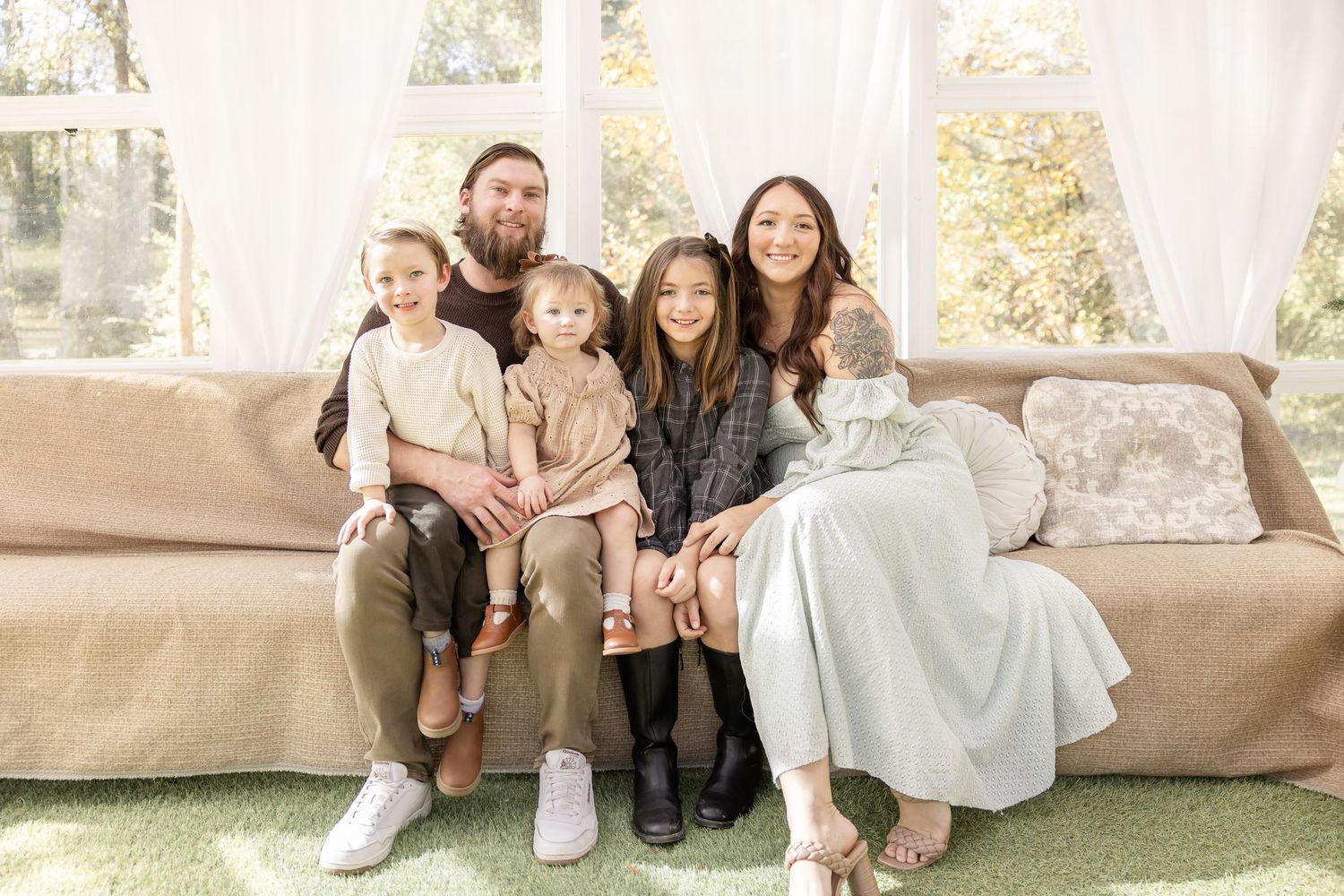A family sits together on a light colored couch while posing for portraits in coordinated outfits.