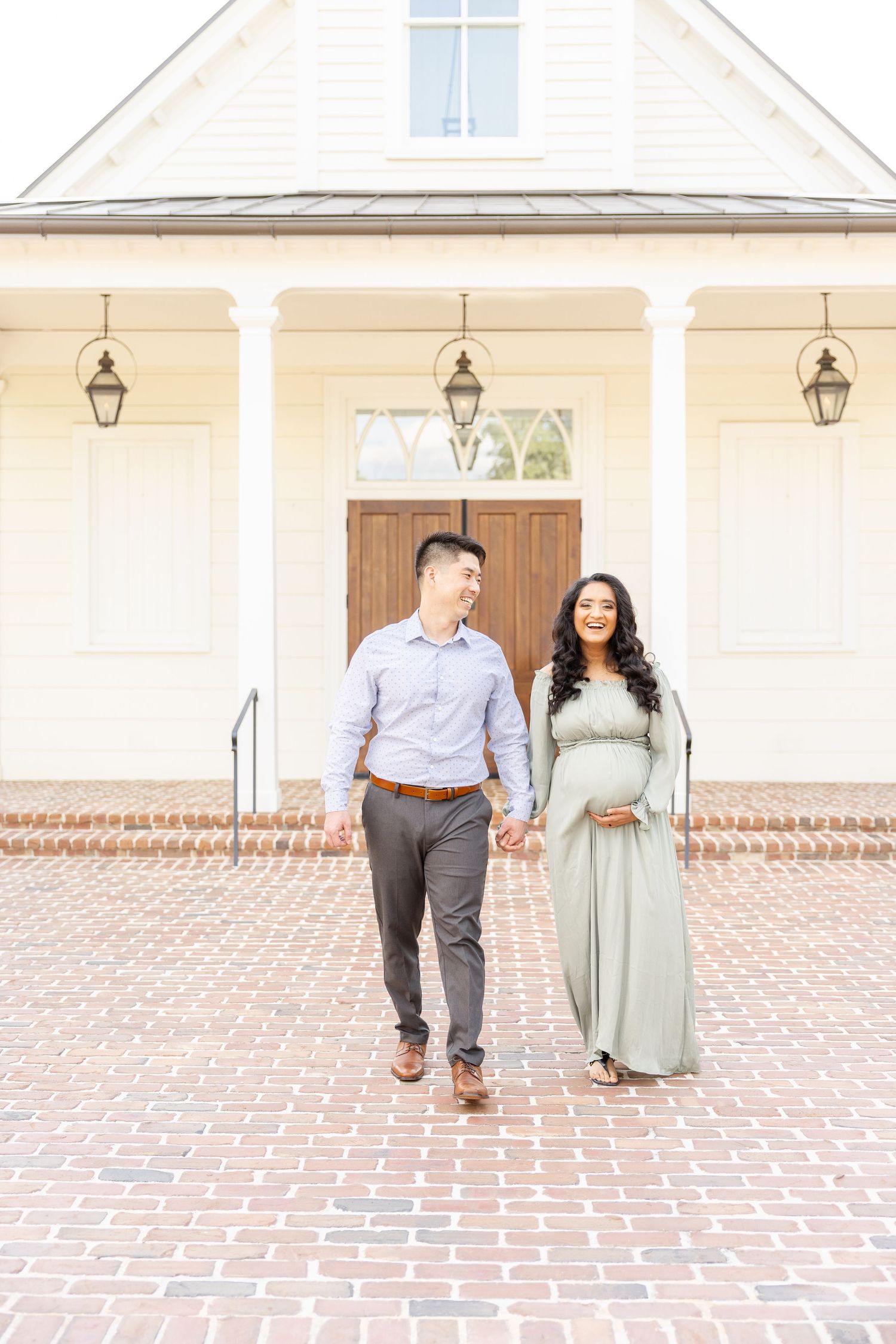 Expecting couple holds hands walking on brick path before white building during their Columbus, GA maternity portrait session.