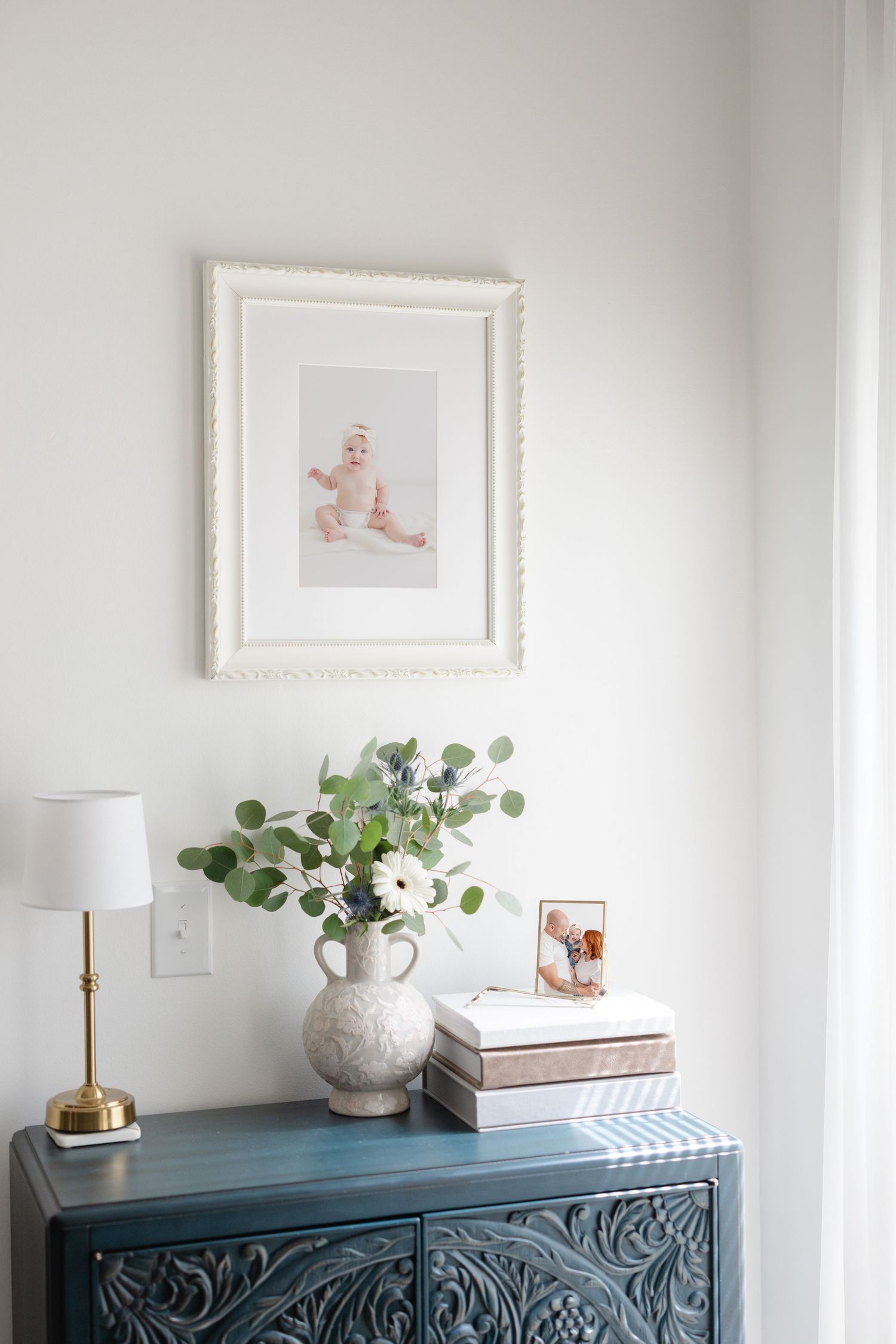 Teal carved dresser with floral arrangement, brass lamp, and framed baby photo above in a bright, minimalist nursery corner.