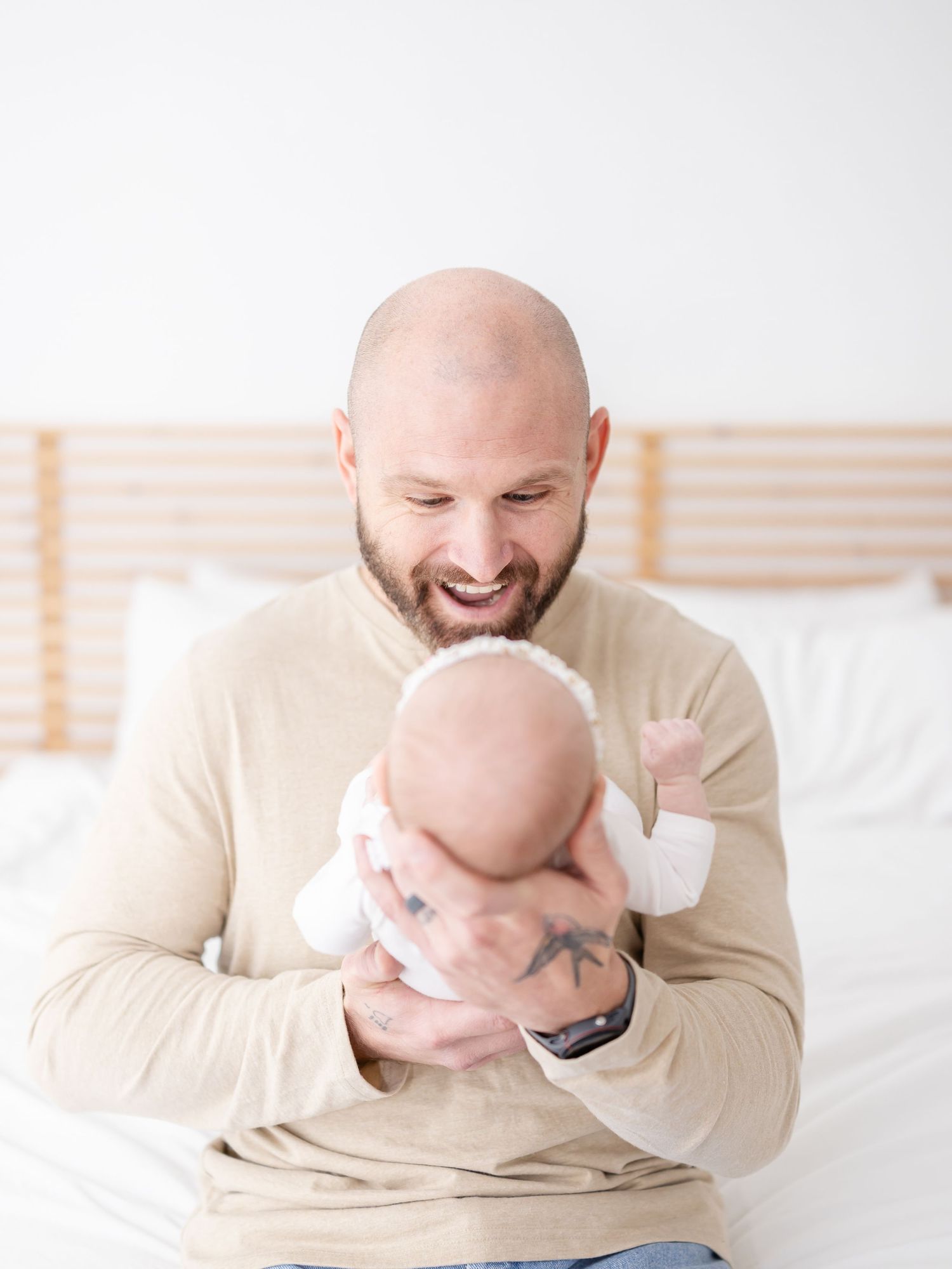 A father in a beige sweater sits on a white bed while holding and smiling at his baby by a Professional newborn photographer in Columbus GA.