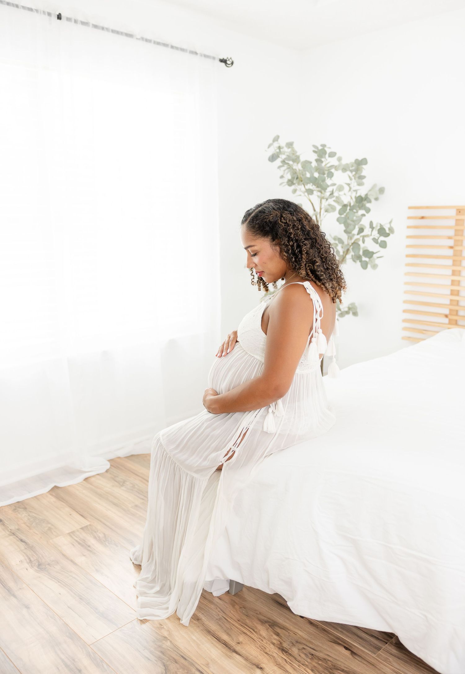Pregnant woman in white dress sitting on bed cradling her baby bump in a bright, minimalist bedroom.