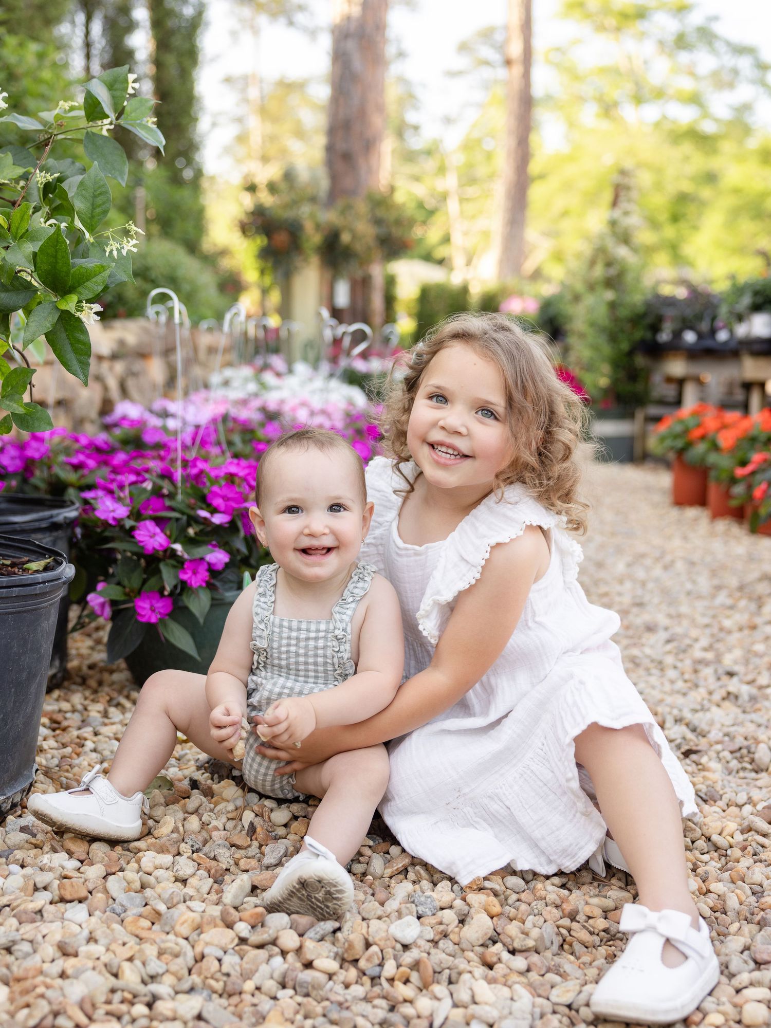 Two young siblings smile together among colorful garden flowers, with the older girl in white hugging her baby brother.