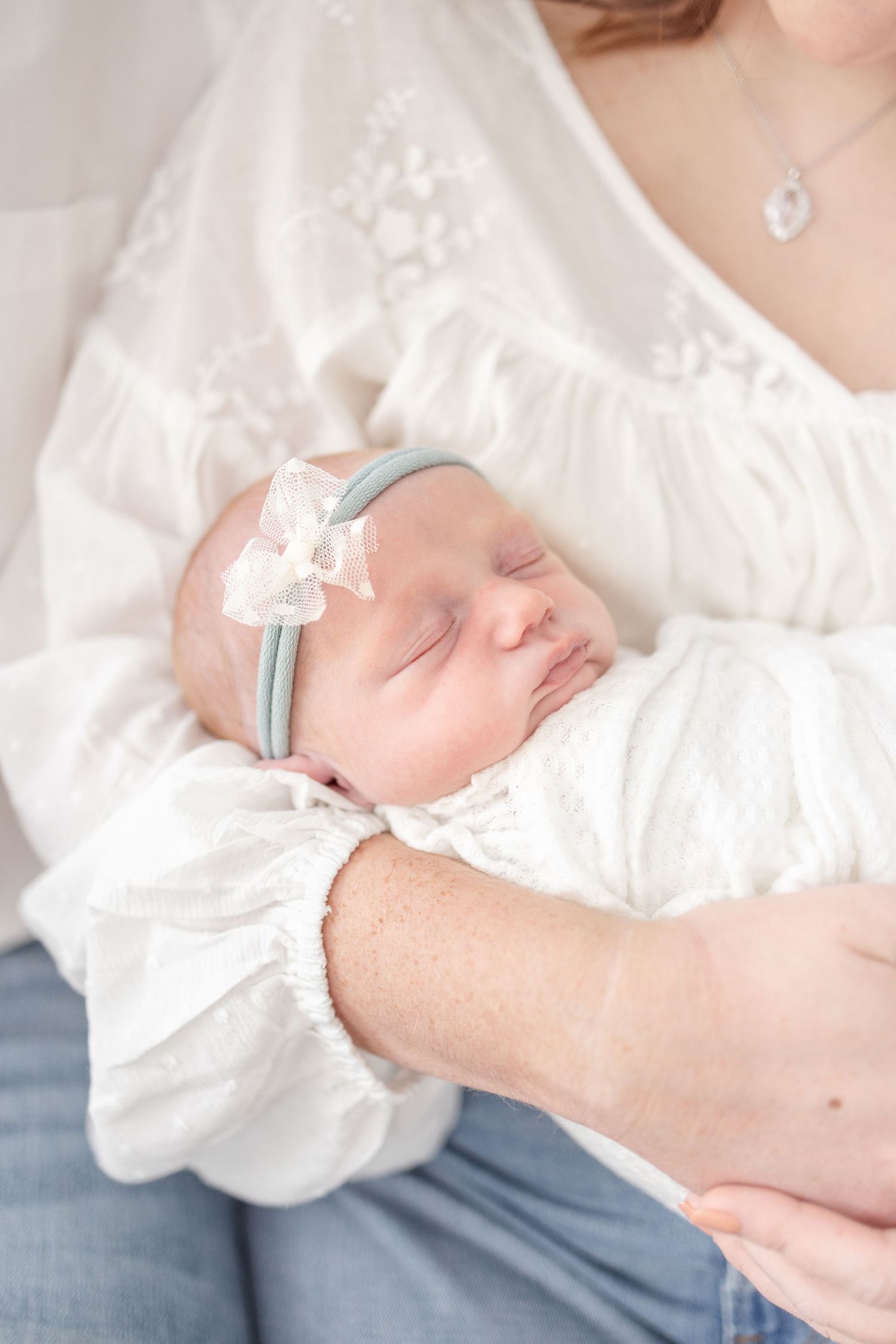 Newborn baby girl sleeping peacefully in white swaddle with pink bow headband, cradled in mother's arms.
