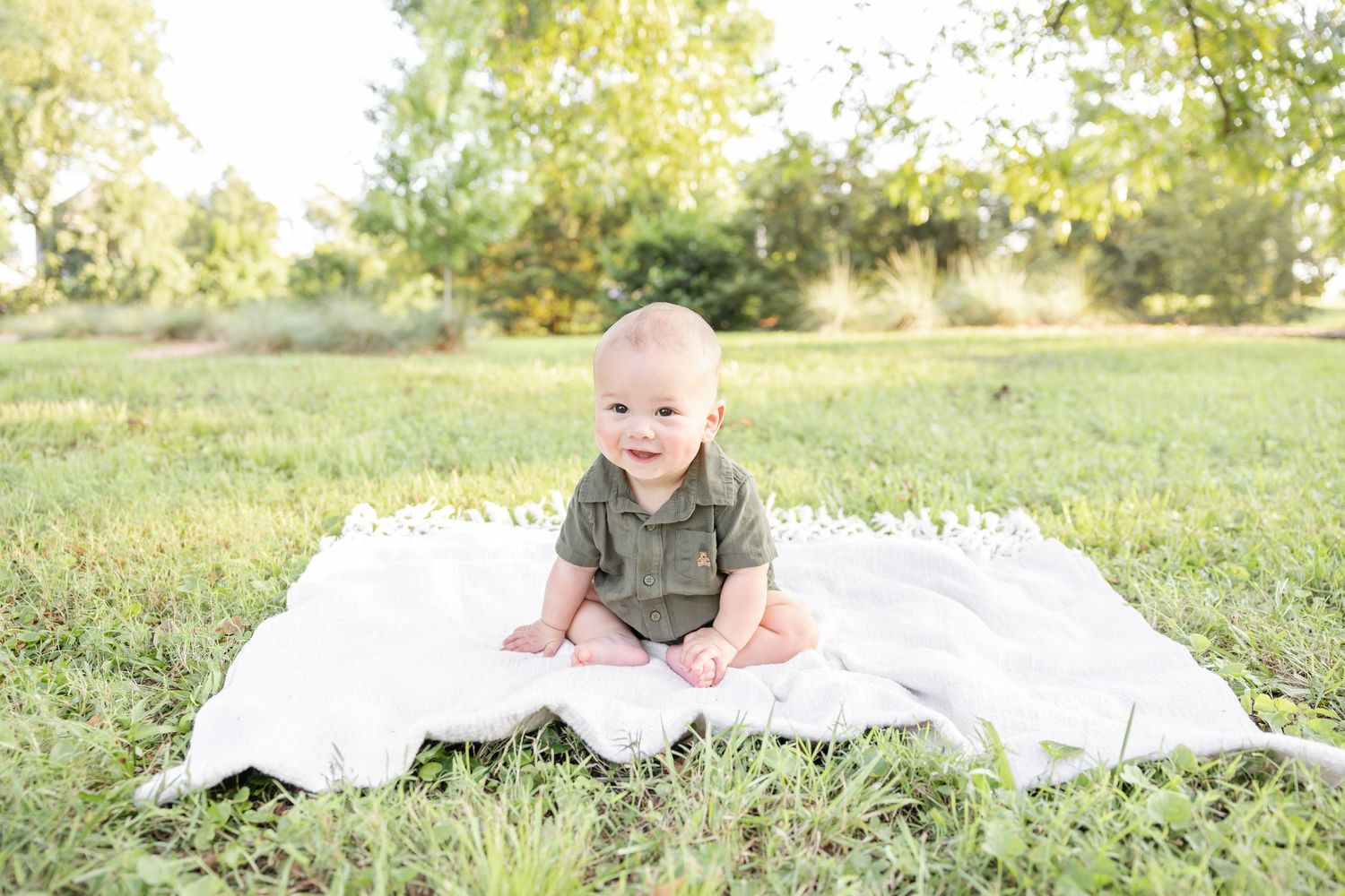A small child wearing a green onesie sits on a white blanket in a sunny grass field on a bright summer day at his 6 month milestone session.