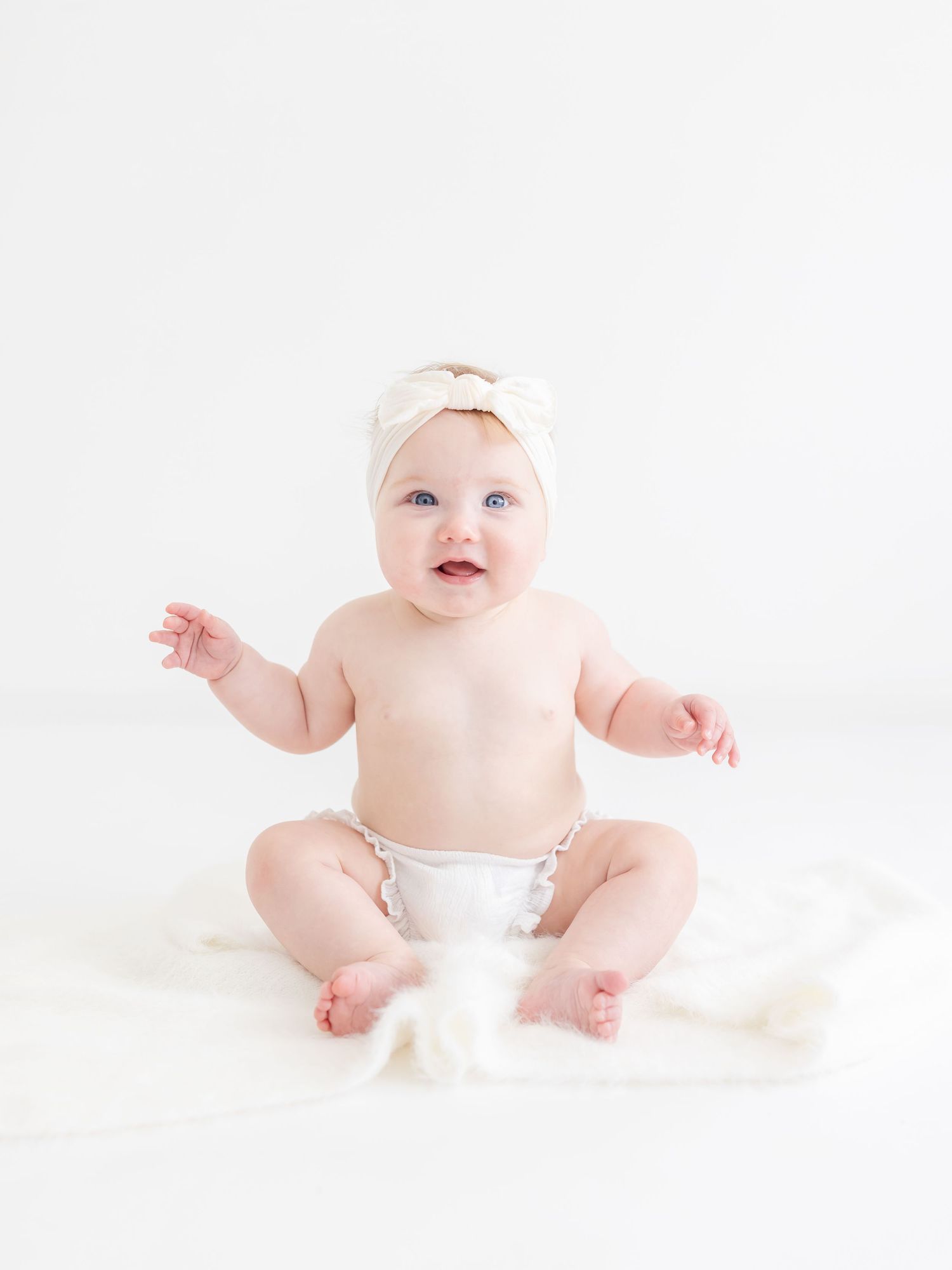 Adorable baby girl sitting upright in a diaper and bow headband on a white fluffy blanket with a bright white background.