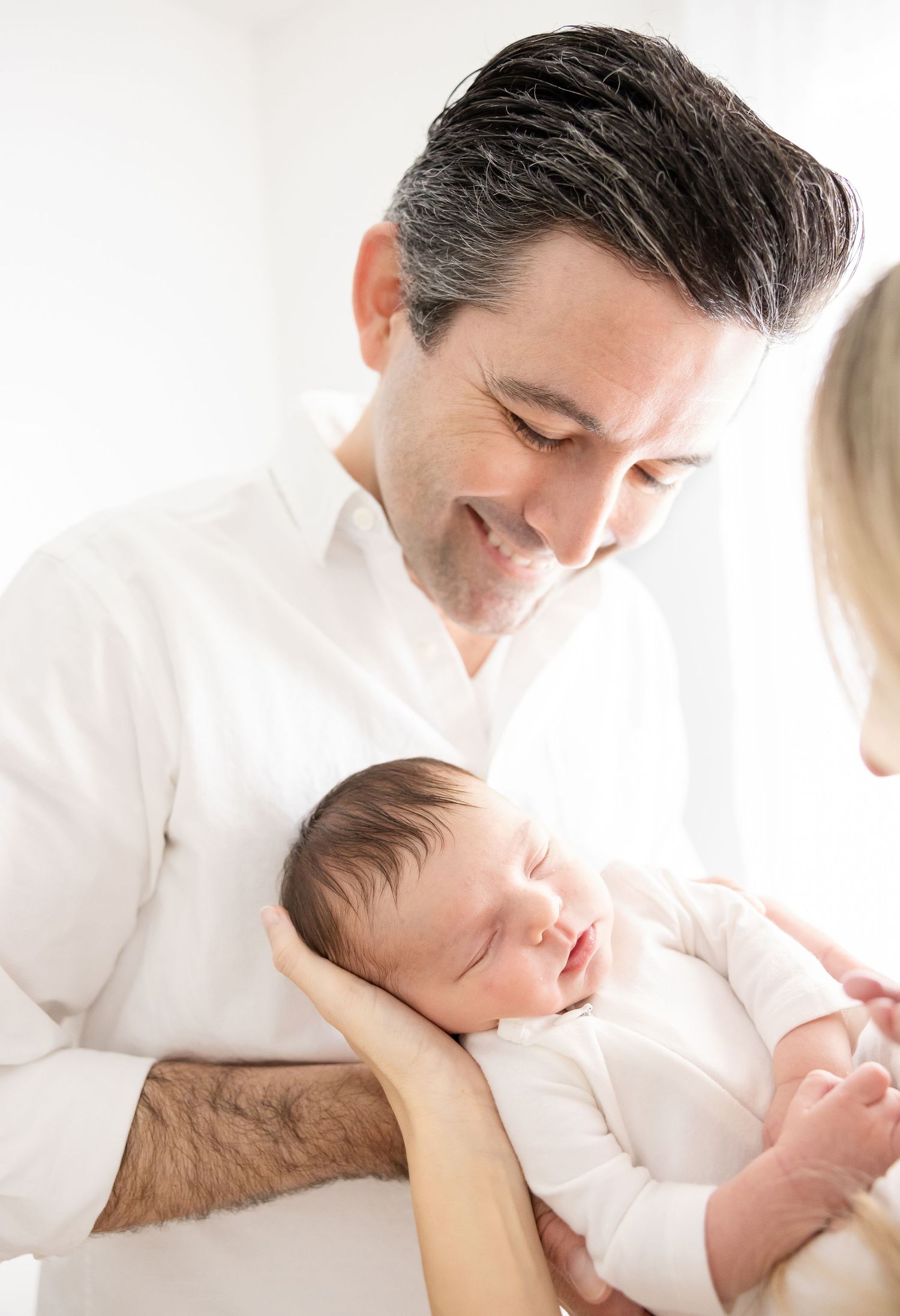 A smiling father in a white shirt tenderly holds his sleeping newborn baby while a woman looks on nearby.