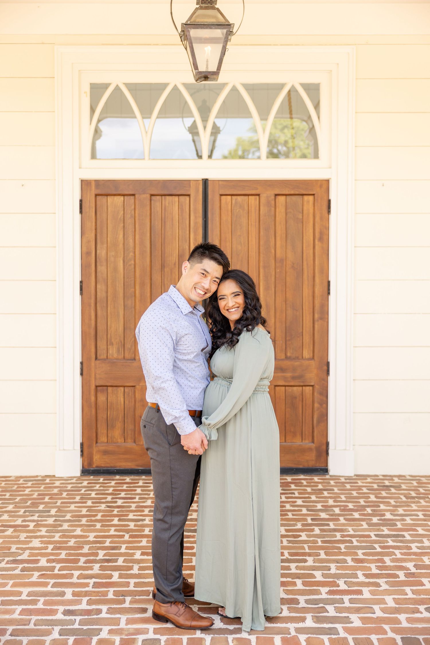 A couple stands together holding hands and smiling on a brick patio in front of wooden church doors at their Columbus, GA maternity session