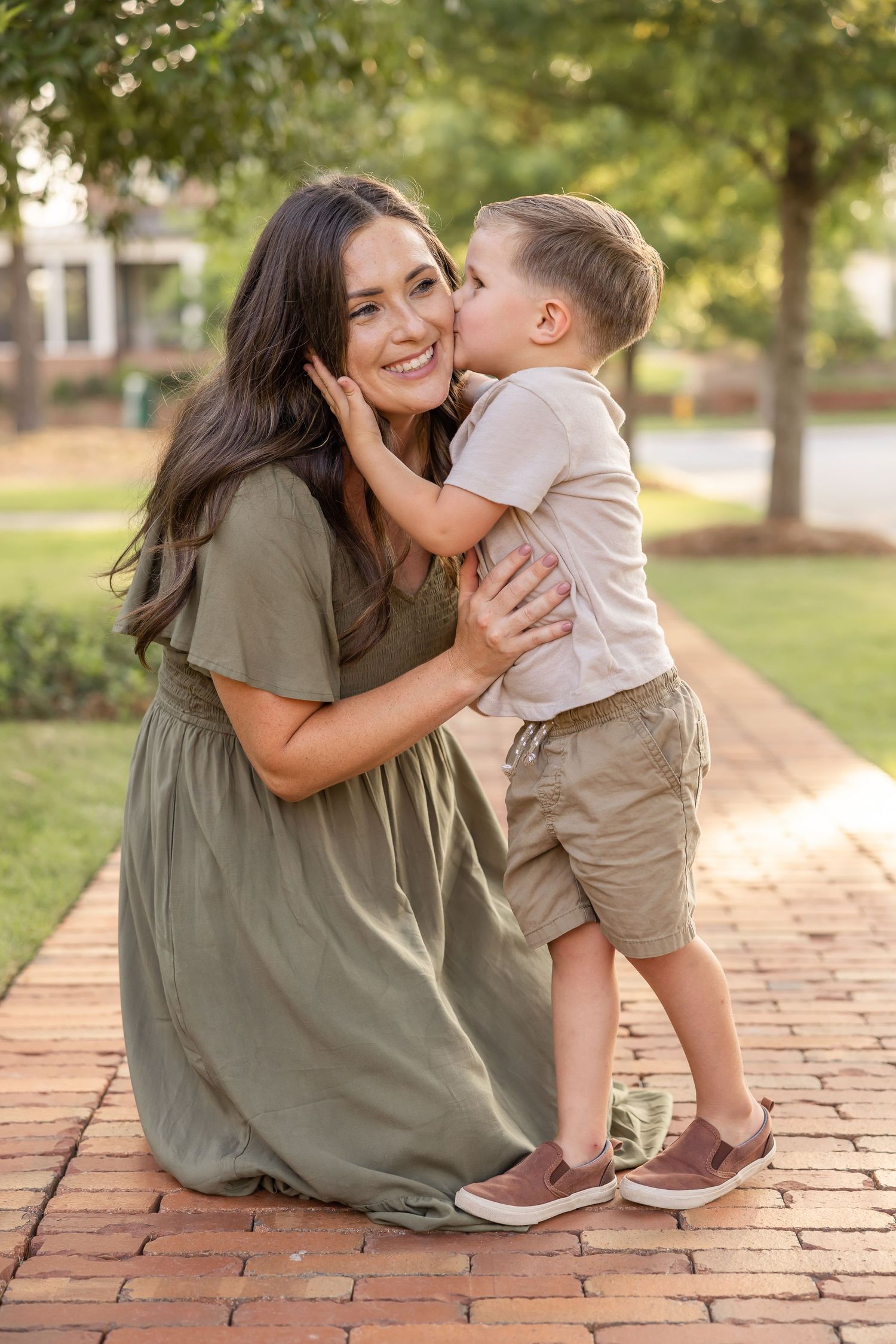 A sweet moment between a mom and her son. The son holds mom's smiling face as she kneels down on the brick sidewalk and kisses her cheek.