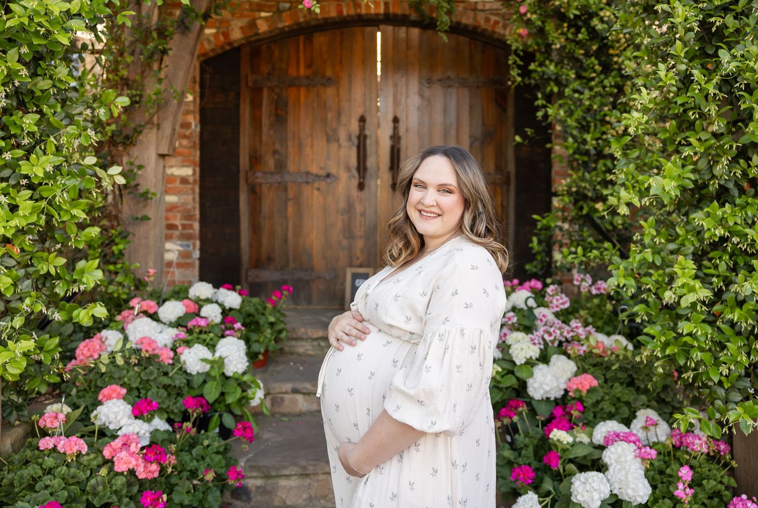 Pregnant woman in floral dress smiling outdoors, surrounded by colorful flowers by a rustic wooden door by Auburn, AL maternity photographer