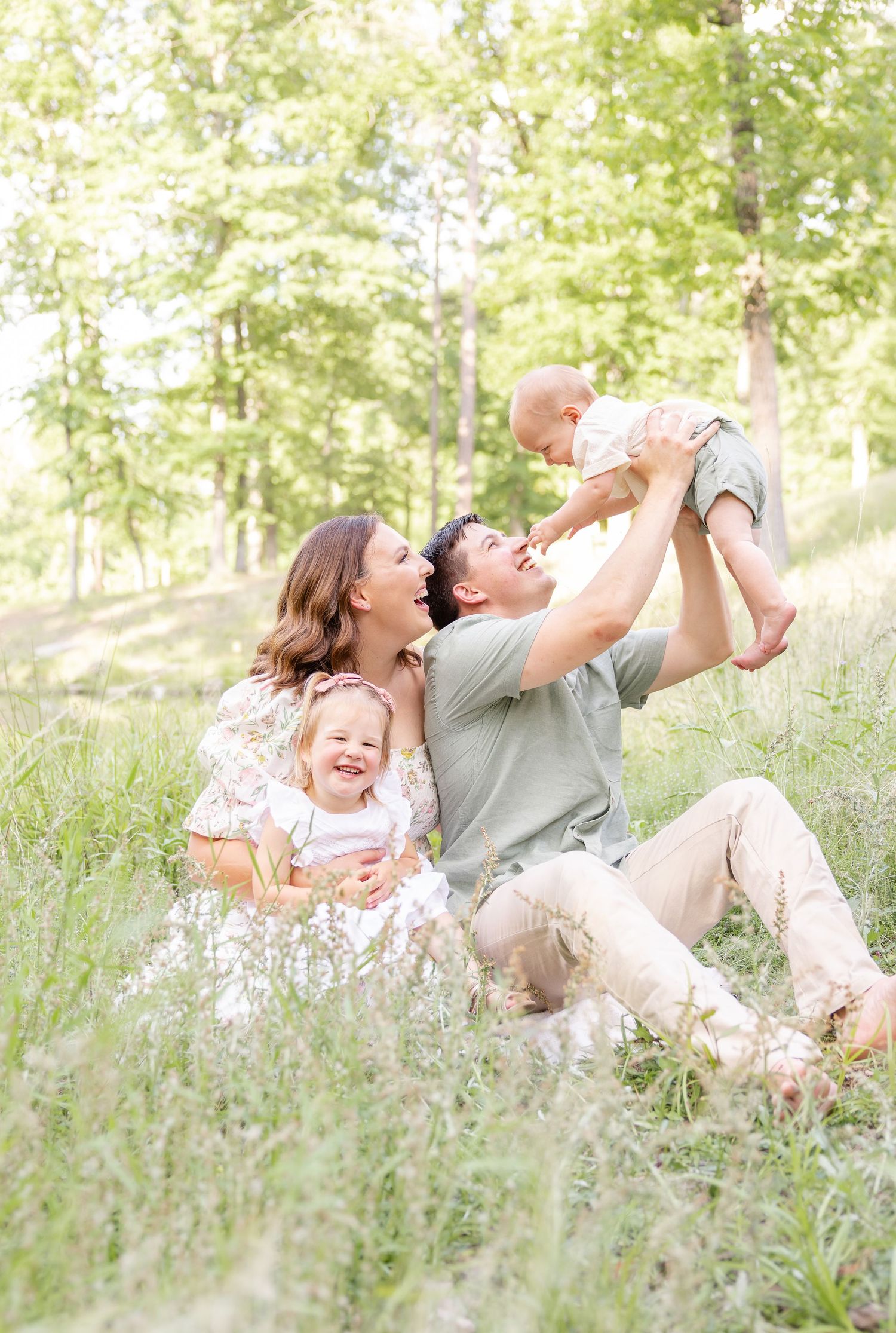 Happy family sitting in green meadow, parents laughing with toddler girl and baby lifted in air during their Columbus, GA spring portraits.