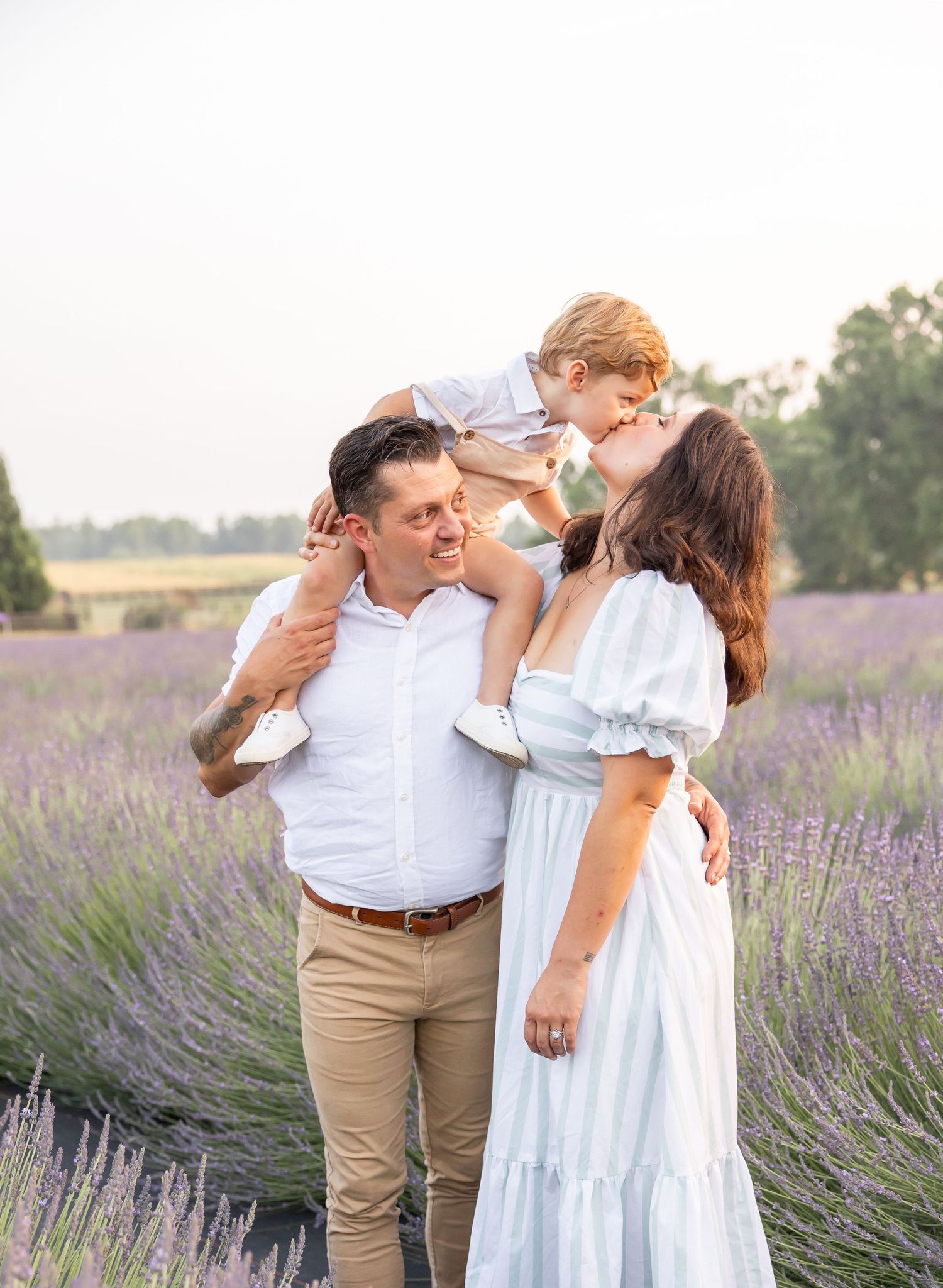 Family of three posing in a lavender field, father carrying son on shoulders while mother in white dress smiles warmly.