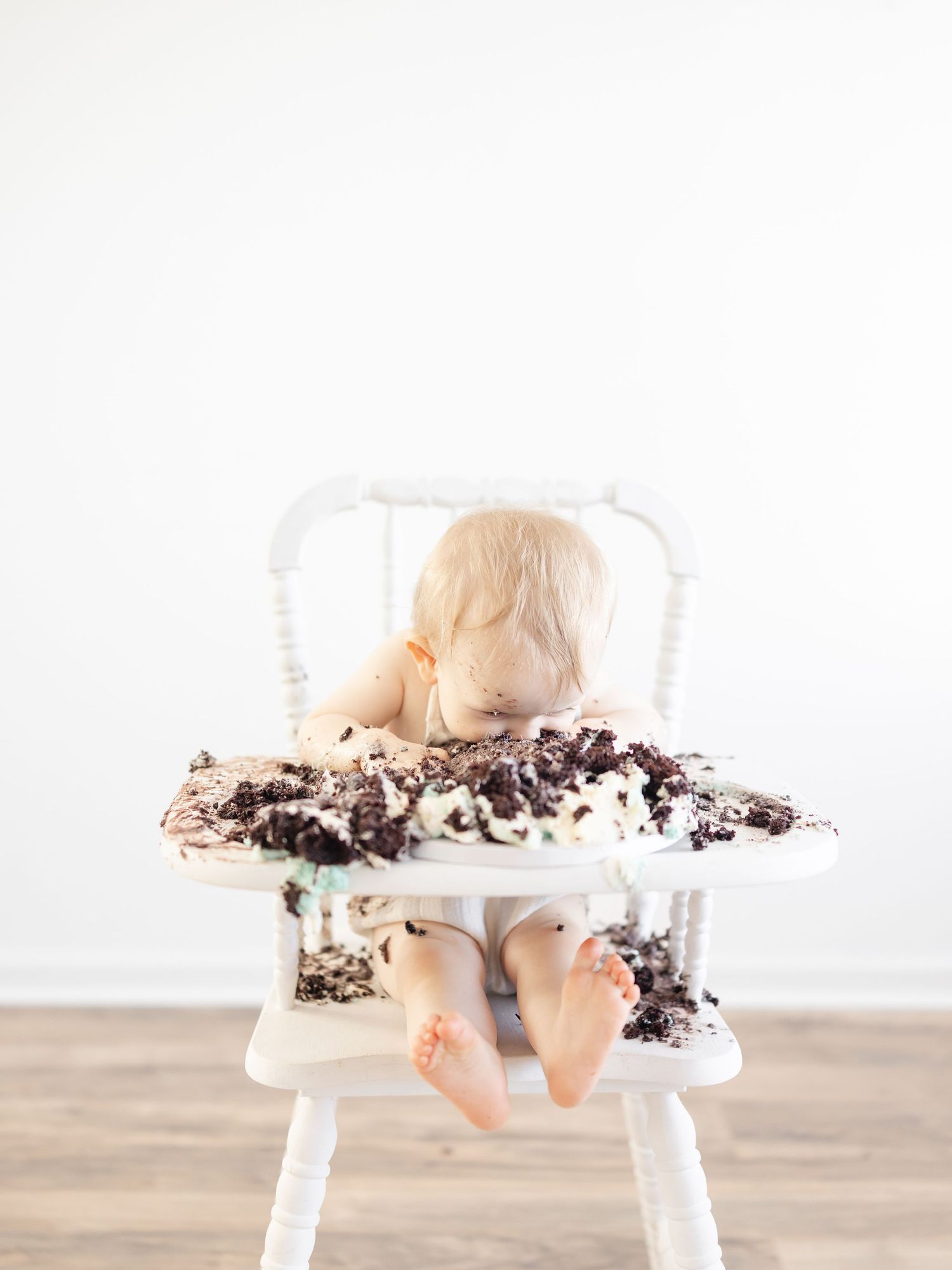 A messy baby sitting in a white high chair covered in chocolate cake during a first birthday smash cake photo session.