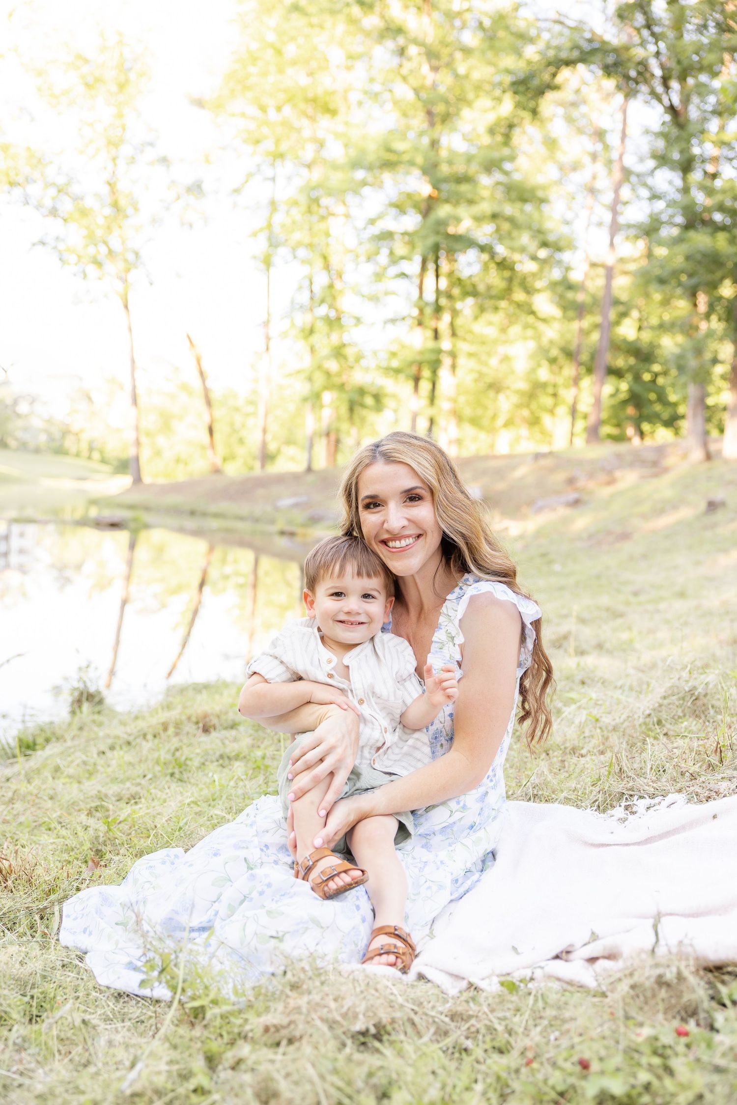 A mother and young toddler sit together on a white blanket near a pond on a sunny summer day.