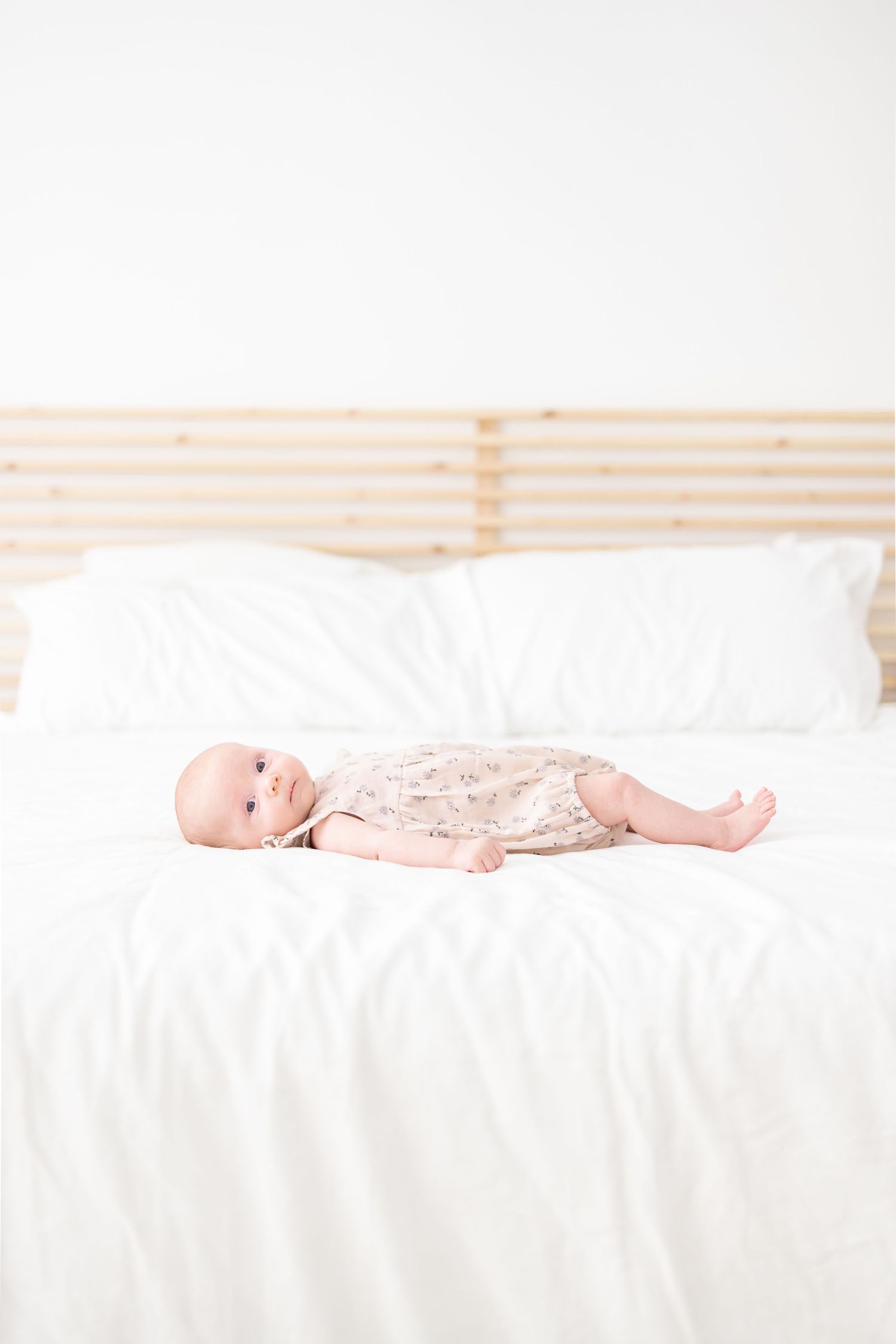 Baby wearing pink lace outfit lying on white bedding with wooden headboard in bright minimalist setting.
