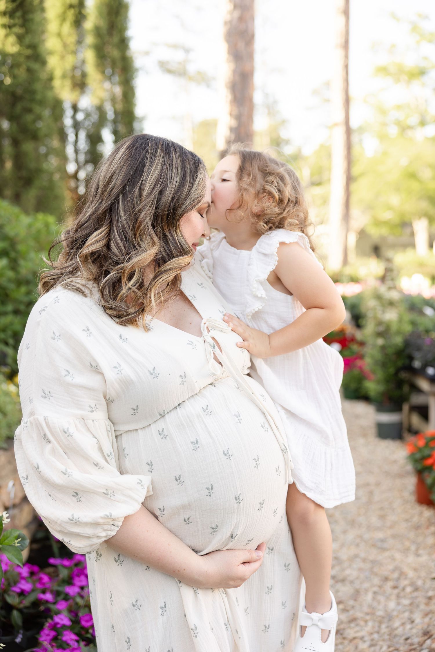 Pregnant woman in white floral dress holds toddler girl sharing a kiss outdoors in a garden setting by Auburn, AL maternity photographer.