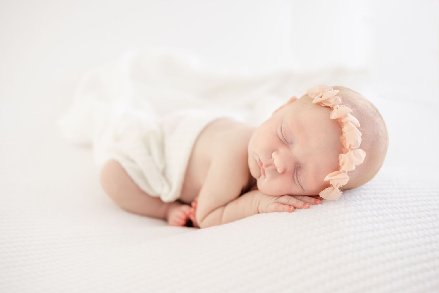 Sleeping newborn baby wearing delicate pink headband lying on soft white blanket in peaceful slumber.