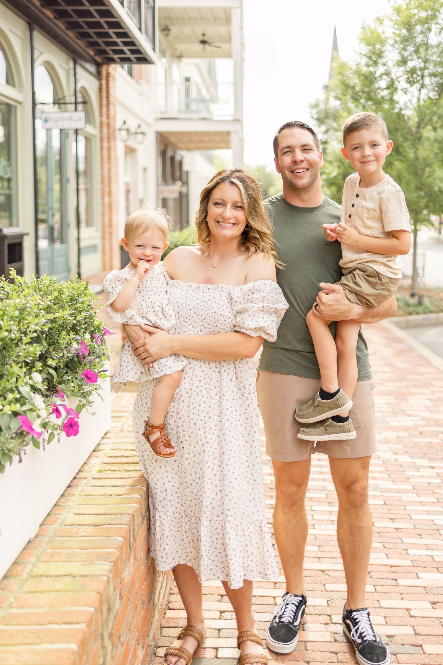 A family stands together on a brick sidewalk outside a storefront building with flowers and greenery.
