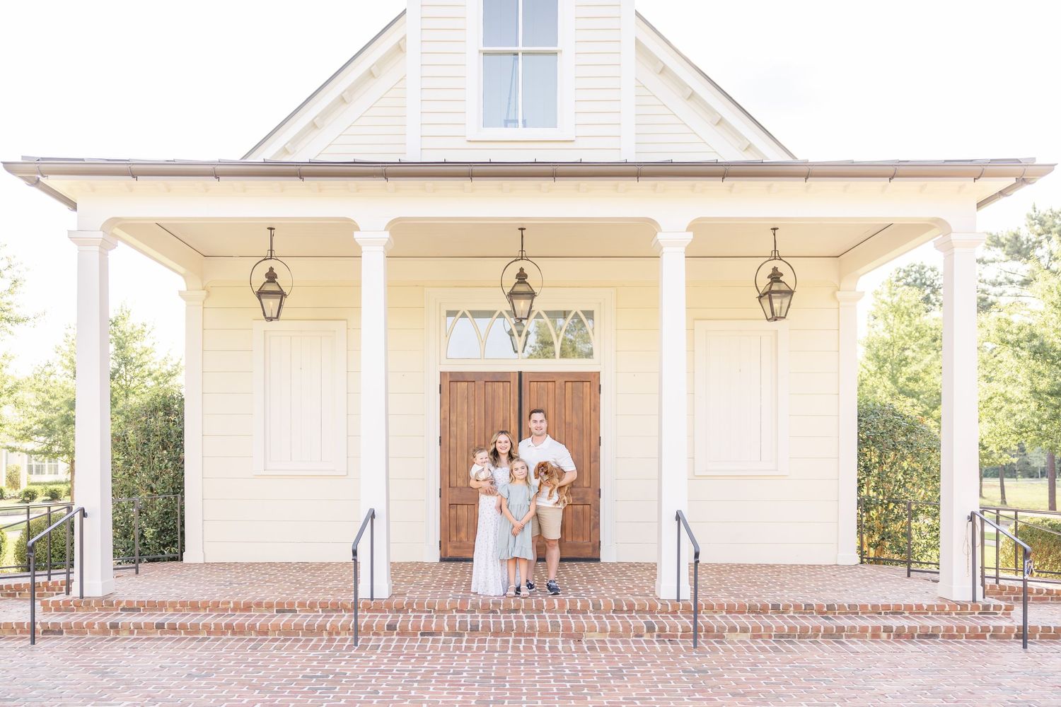 Family posing in front of a charming white chapel with brick steps and lanterns.